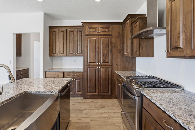 3636 West 910 South Syracuse, UT 84075 - Photo 36 of 70 Kitchen featuring wood-finish cabinetry, light-toned granite countertops, a stainless steel farmhouse sink, a stainless steel gas range with a matching range hood, and wood-finish flooring