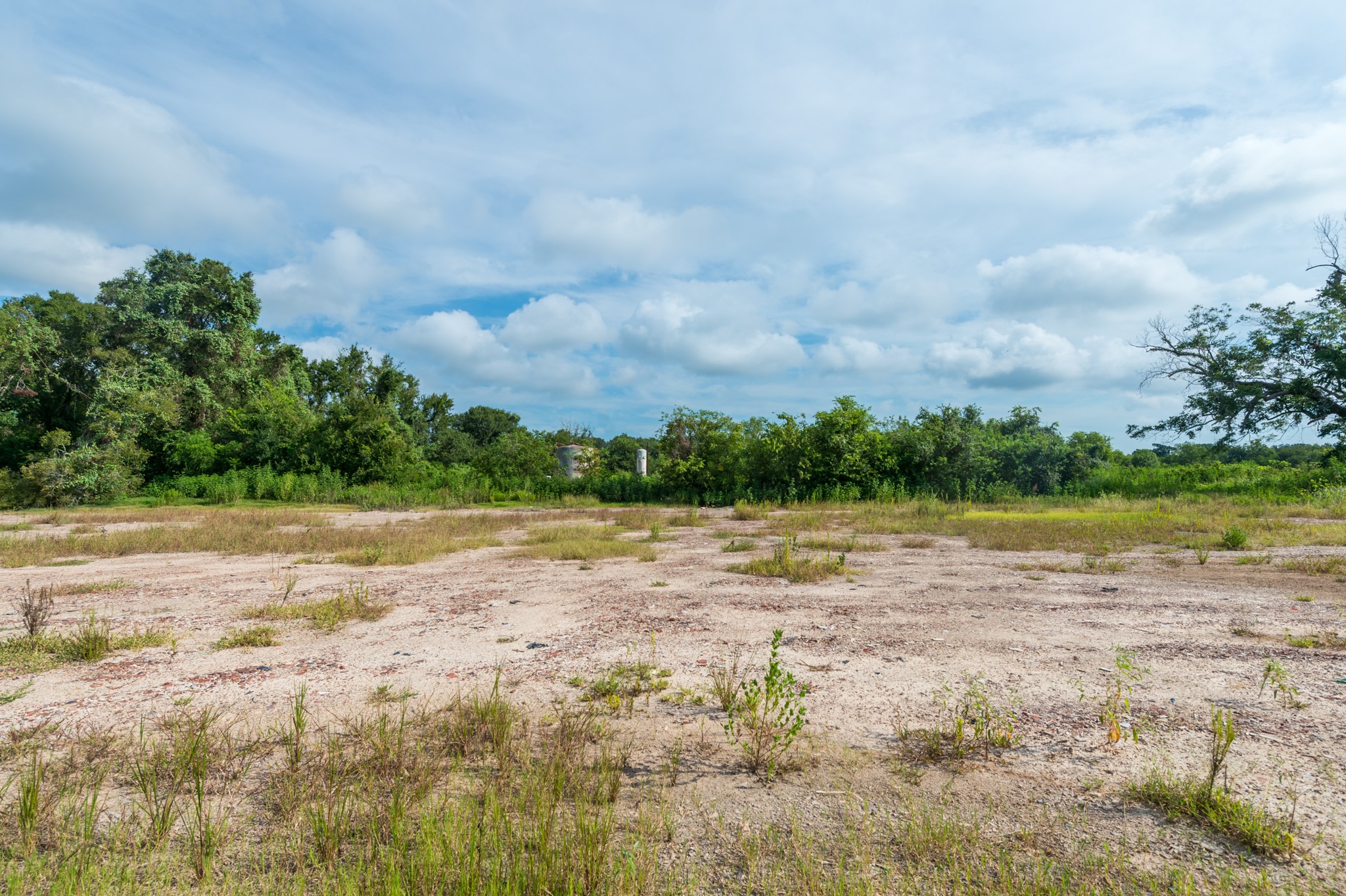1353 Seidel Road Brookshire, TX 77423 - Photo 15 of 35 This photo shows a spacious, open plot of land with sparse vegetation and surrounded by lush greenery, under a partly cloudy sky. Ideal for building a home with a natural setting.