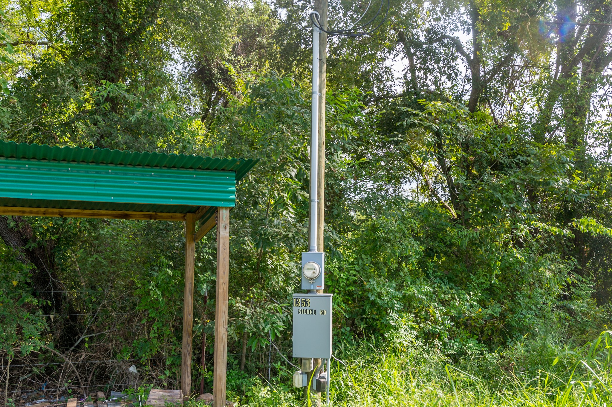 1353 Seidel Road Brookshire, TX 77423 - Photo 17 of 35 This photo shows a utility pole with a meter and a small green-roofed structure, set against a backdrop of dense, leafy trees.