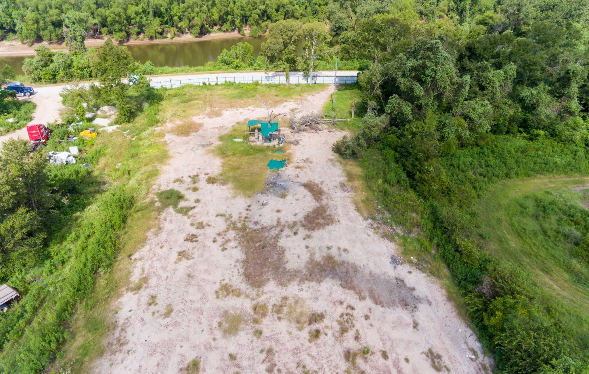1353 Seidel Road Brookshire, TX 77423 - Photo 25 of 35 This photo shows a spacious, partially cleared lot with a mix of grassy and sandy areas, surrounded by trees. A road runs along one edge, and there's a view of a river in the distance, providing a natural and serene setting.