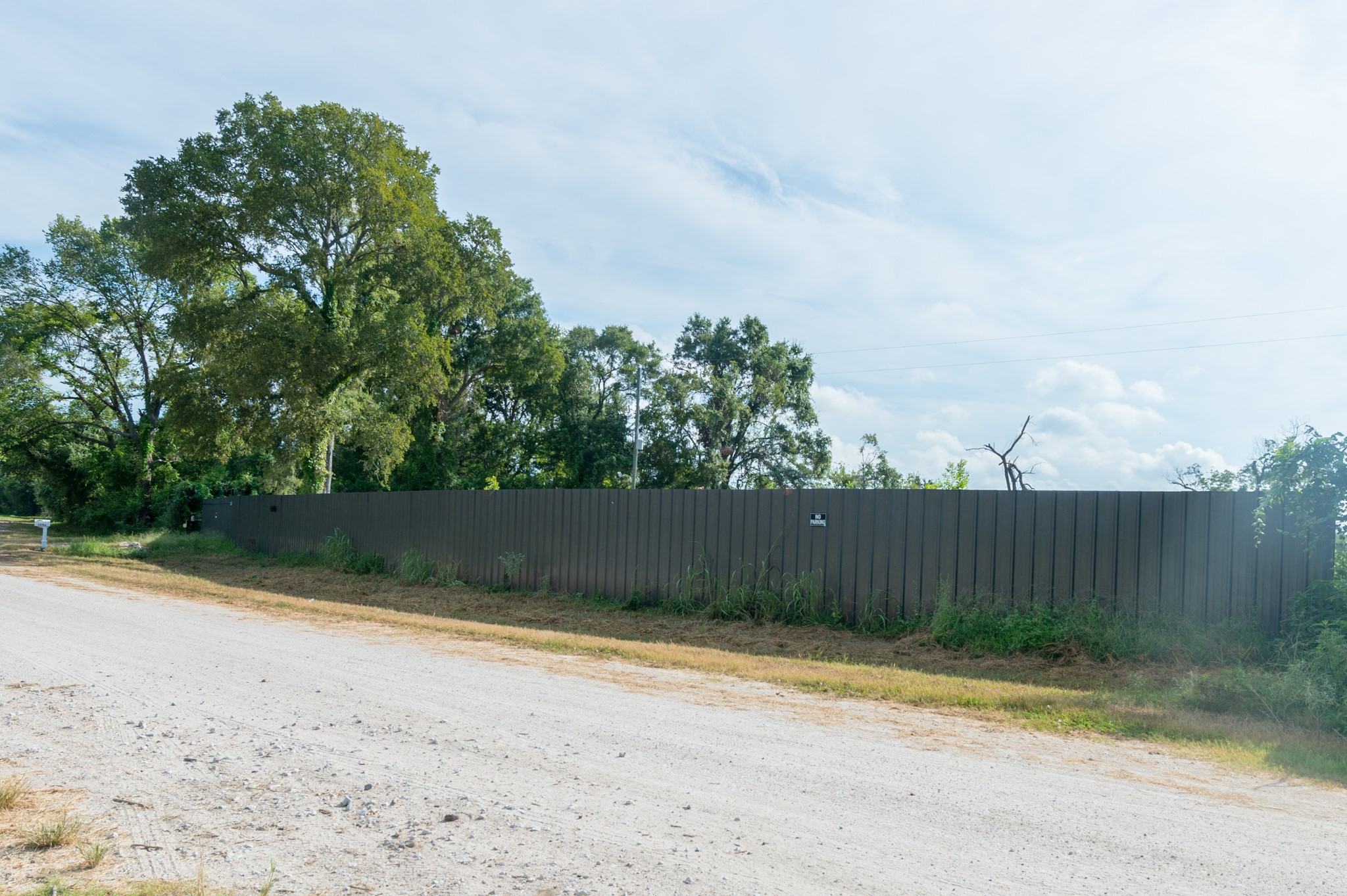1353 Seidel Road Brookshire, TX 77423 - Photo 6 of 35 A long metal fence lines a dirt road, bordered by lush trees and greenery, providing privacy and a natural backdrop.