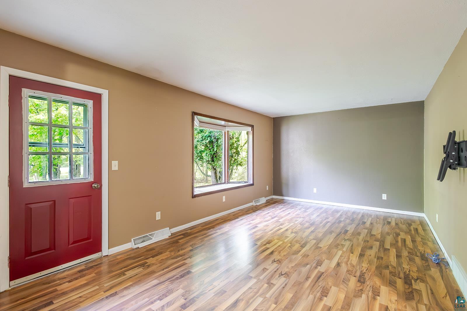 7045 Morris Thomas Road Cloquet, MN 55720 - Photo 9 of 28 Expansive room featuring wood-finish flooring, a red entry door with a multi-pane glass insert, a wide bay window, and a wall-mounted bracket