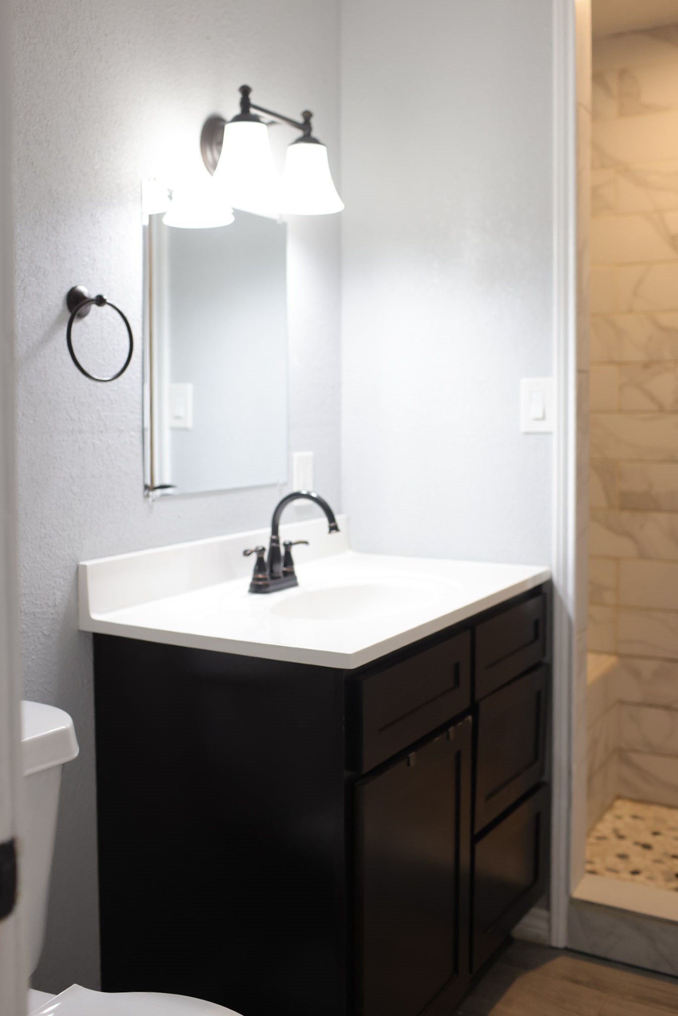 407 Tamarind Street Lake Jackson, TX 77566 - Photo 29 of 36 This photo shows a modern bathroom with a sleek dark vanity and white countertop. It features a mirror with dual overhead lighting and a glimpse into a tiled shower area.