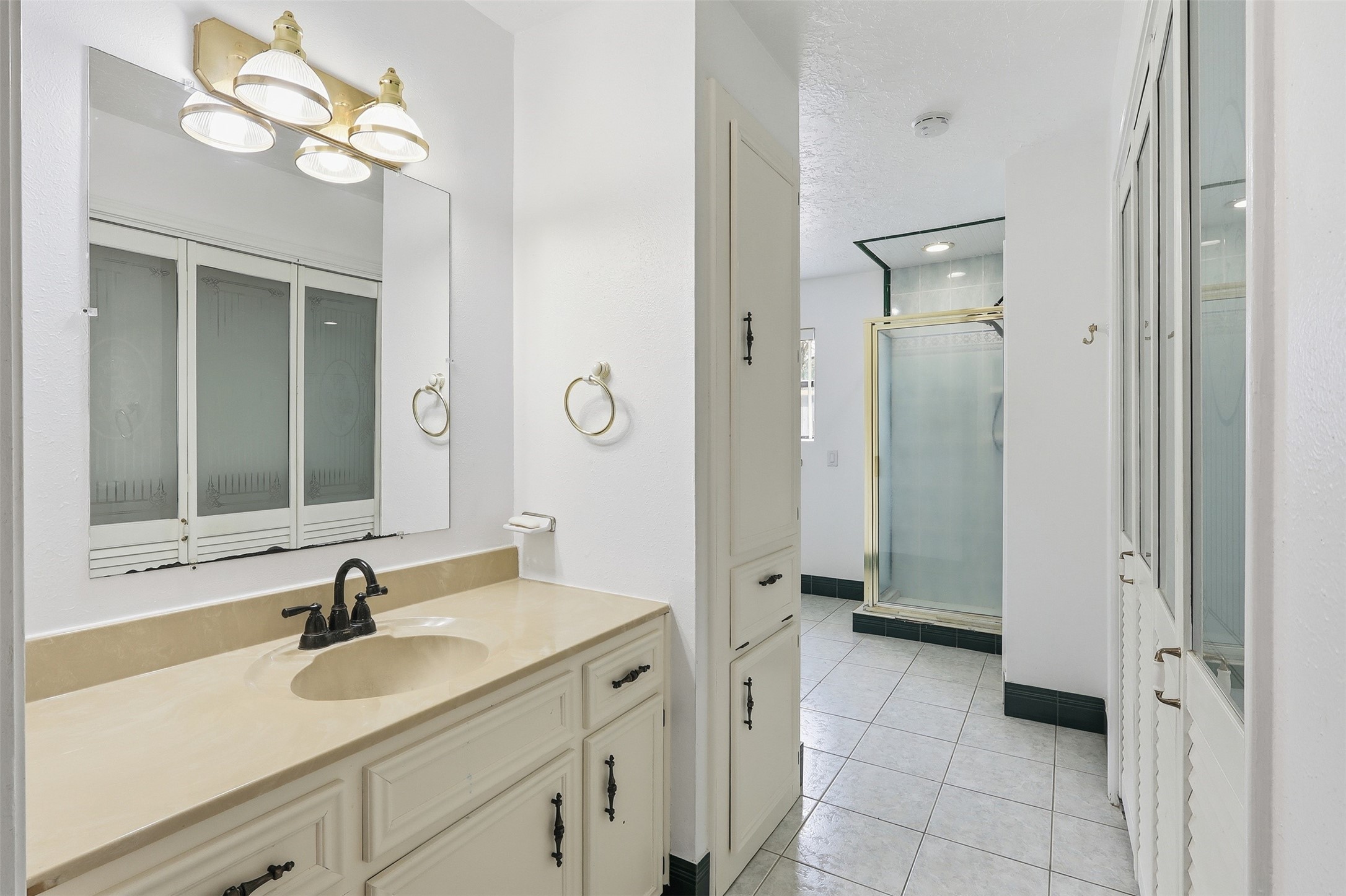 614 Tallow Drive Conroe, TX 77385 - Photo 22 of 36 This bathroom features a spacious vanity with a large mirror and three modern light fixtures. It includes ample storage with cabinets and a walk-in shower with a glass door. The white tile flooring and walls give it a bright, clean look.