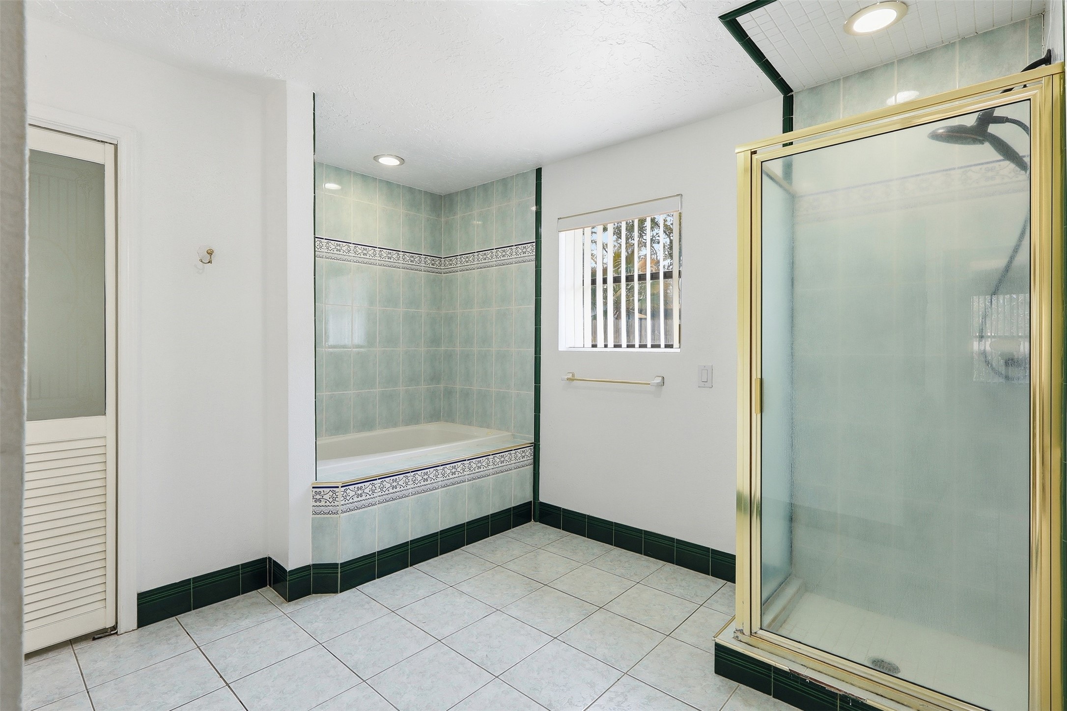 614 Tallow Drive Conroe, TX 77385 - Photo 24 of 36 Bright bathroom featuring a spacious glass shower, a tiled bathtub, and a window with blinds. The room is adorned with light and dark green tiles, creating a fresh and clean look.