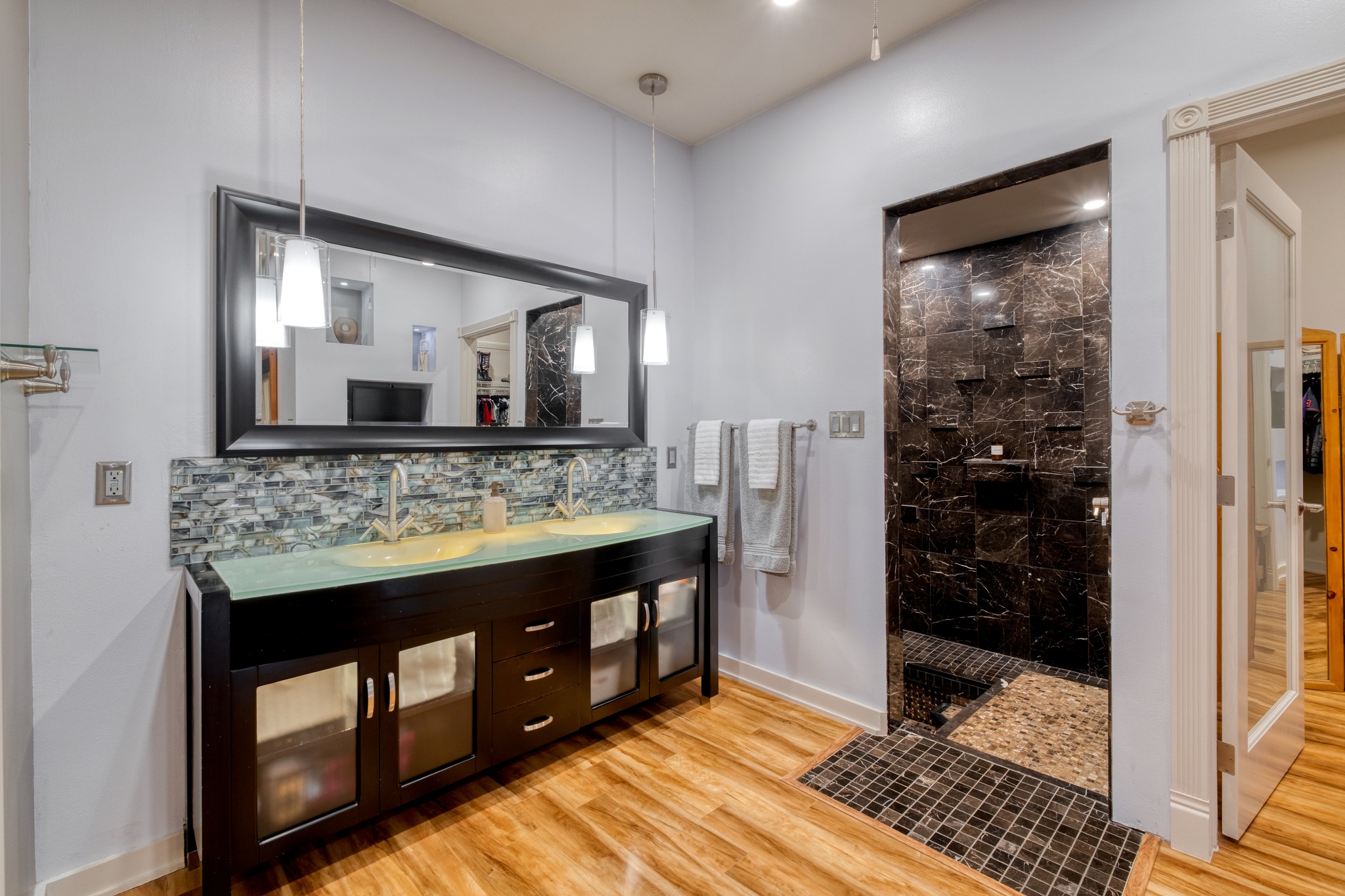 1474 County Rd B Lexington, TX 78947 - Photo 8 of 34 A refined bathroom featuring a dual-sink vanity with clear glass countertops and a mosaic tile backsplash, complemented by a large, frameless shower enclosure.