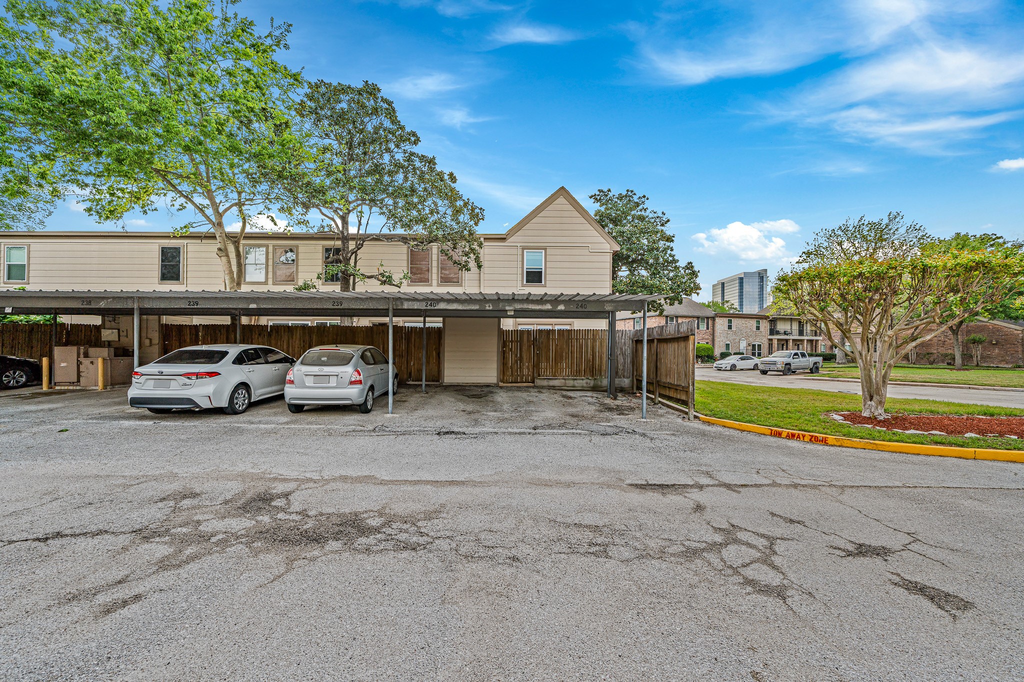830 Threadneedle Street, Unit 240 Houston, TX 77079 - Photo 28 of 33 This photo shows a townhouse with a covered carport for two vehicles, surrounded by mature trees and a well-maintained lawn. The area appears to be part of a residential neighborhood with nearby buildings visible in the background.