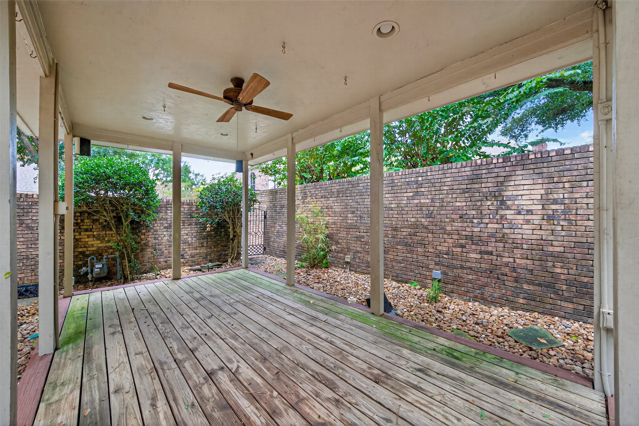 901 Longmire Road, Unit 13 Conroe, TX 77304 - Photo 46 of 46 This photo features a covered patio with a wooden deck and ceiling fan, surrounded by a brick wall for privacy. It's an inviting outdoor space with some greenery, perfect for relaxation or entertaining.