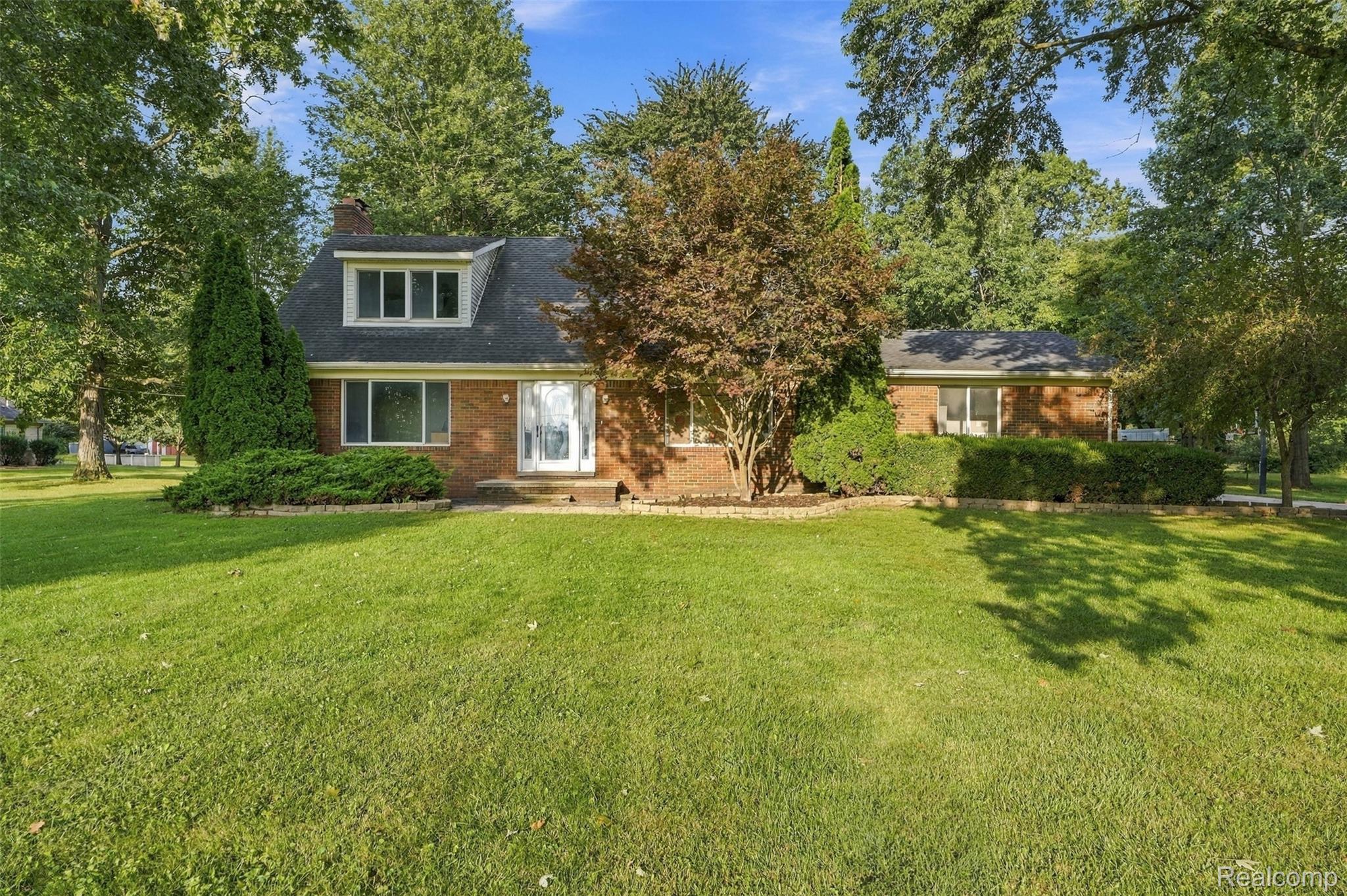 Brick exterior with dormer window, mature trees, established landscaping, and a spacious lawn
