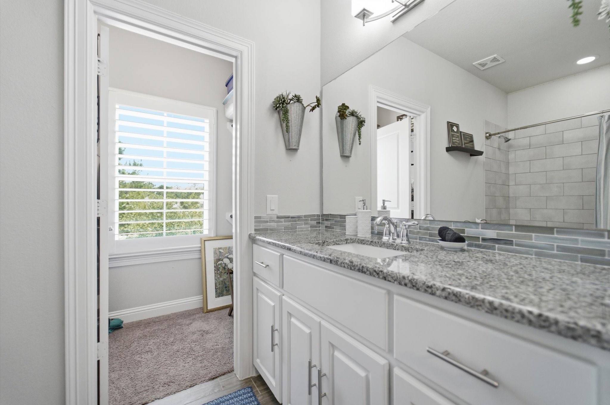 23722 Thortons Park Lane Spring, TX 77389 - Photo 33 of 48 This bathroom features a modern design with a double vanity and granite countertops. It has a large mirror, stylish light fixtures, and a tiled shower. Natural light streams in through a window with shutters, and the neutral color palette creates a clean, inviting atmosphere.