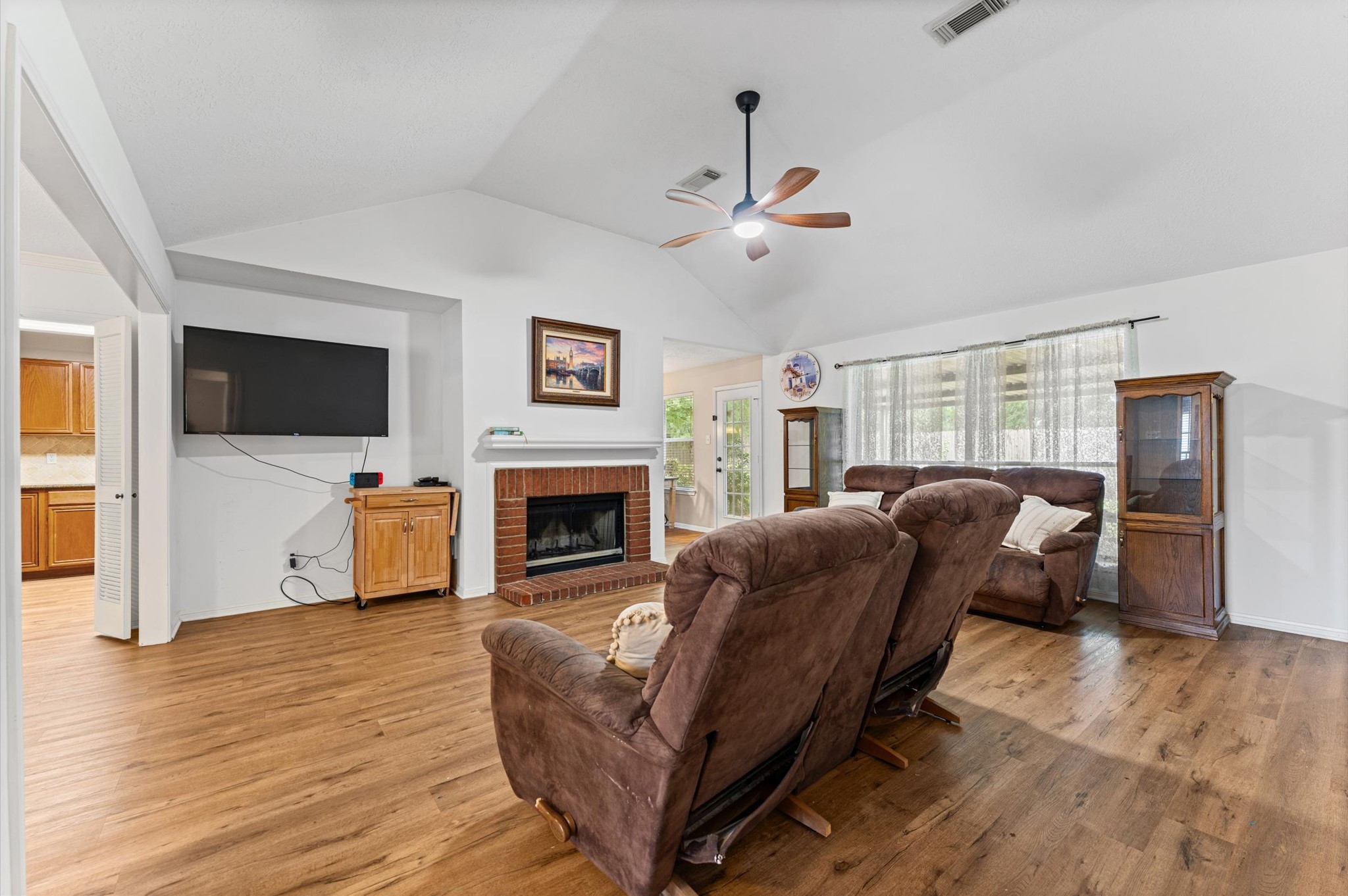 17618 Mill Springs Drive Spring, TX 77379 - Photo 14 of 26 Spacious living room with vaulted ceiling, laminate floors, and ample natural light. Features a brick fireplace, wall-mounted TV, and cozy seating. Open layout connects to the kitchen area. Ideal for comfortable family living.