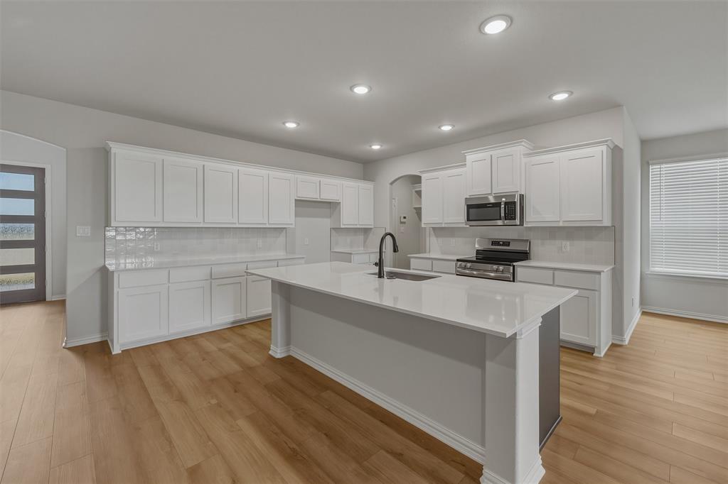 1325 Frio Drive Springtown, TX 76082 - Photo 9 of 24 Kitchen featuring an expansive island with a built-in sink and dark-finish faucet, extensive white cabinetry, light-toned wood-finish flooring, recessed lighting, and stainless steel appliances