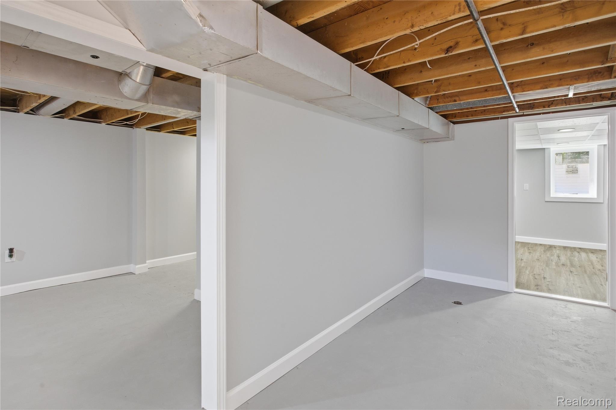 37257 Dundee Street Sterling Heights, MI 48310 - Photo 27 of 47 Unfinished basement space featuring exposed wood joists and ductwork, painted concrete flooring, light gray wall finishes, white trim, and an open doorway leading to an additional finished area with wood-finish flooring and a window