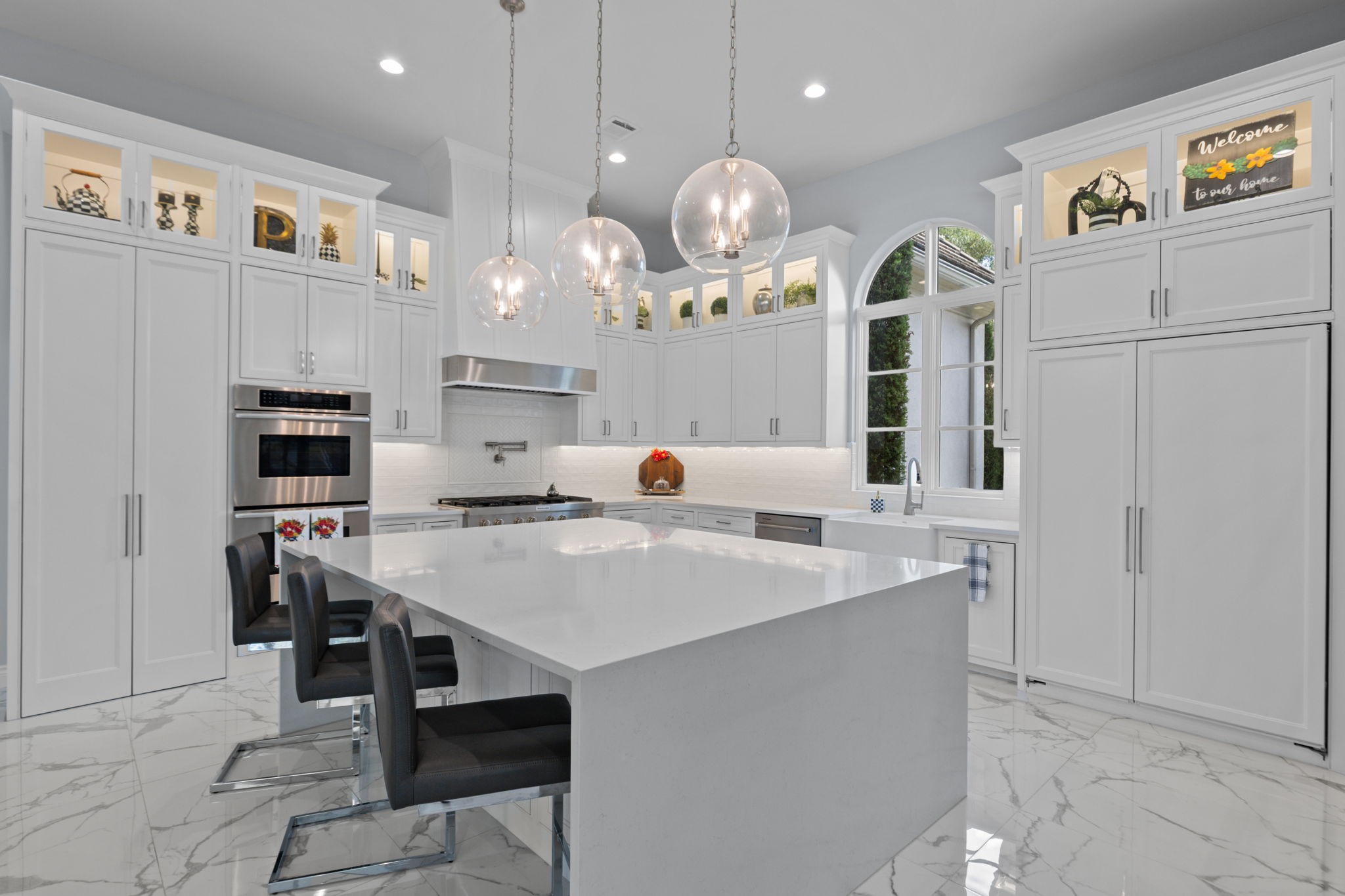 39 Maymont Way Spring, TX 77382 - Photo 13 of 50 This kitchen features a spacious, modern design with sleek white cabinetry, a large island with seating, stainless steel appliances, and elegant pendant lighting. The arched window provides ample natural light, enhancing the bright and airy feel.
