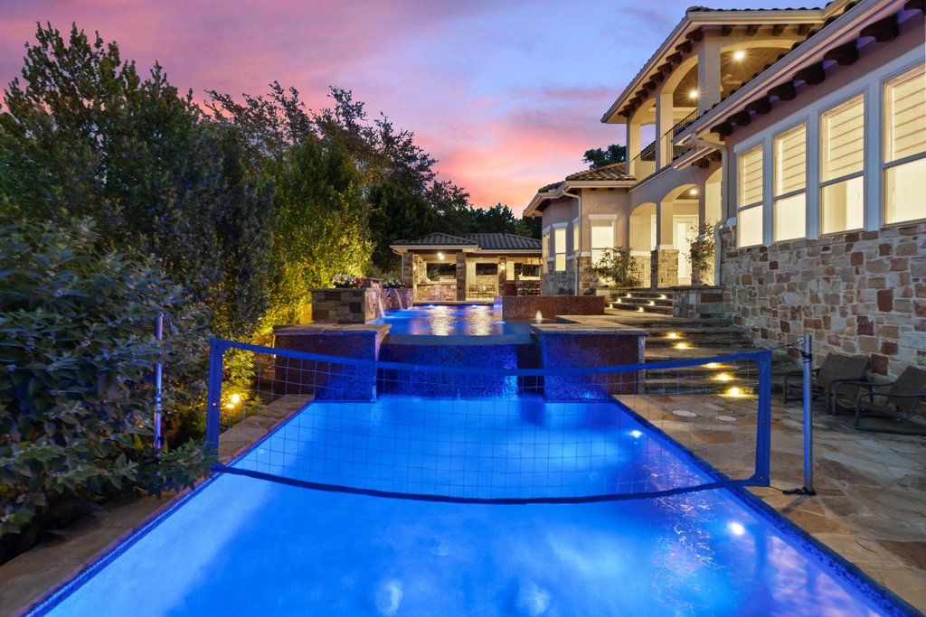 109 Largo Cove Austin, TX 78734 - Photo 31 of 40 Lush landscaping and a sparkling pool with a volleyball net await, alongside a spacious outdoor kitchen and stately stone architecture.