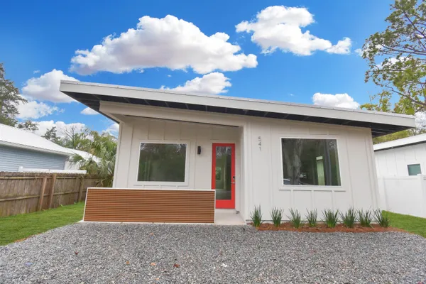 a view of a white house in front of a big yard with plants and large trees