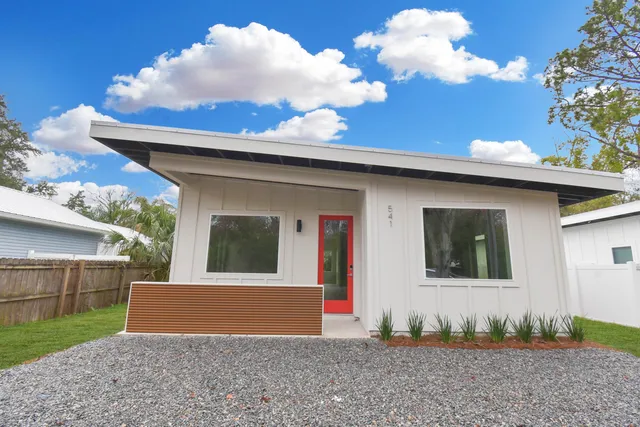 a view of a white house in front of a big yard with plants and large trees
