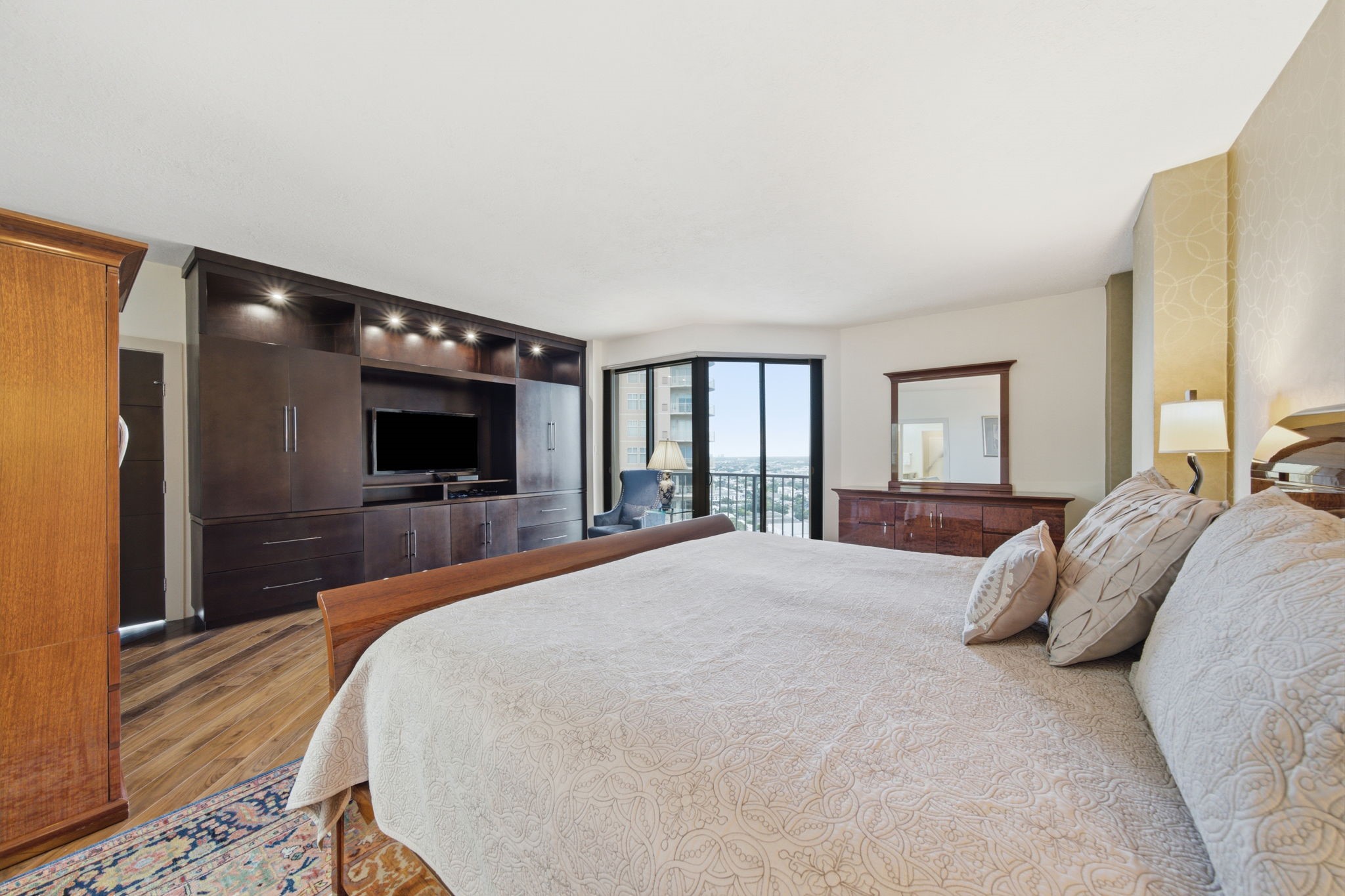 3350 McCue Road, Unit 2402 Houston, TX 77056 - Photo 4 of 49 Custom built-in cabinetry lines the wall of the expansive primary bedroom, providing a dedicated media center and abundant storage alongside rich hardwood flooring. Floor-to-ceiling glass doors open directly to a private balcony, framing elevated city vistas for a serene personal retreat.