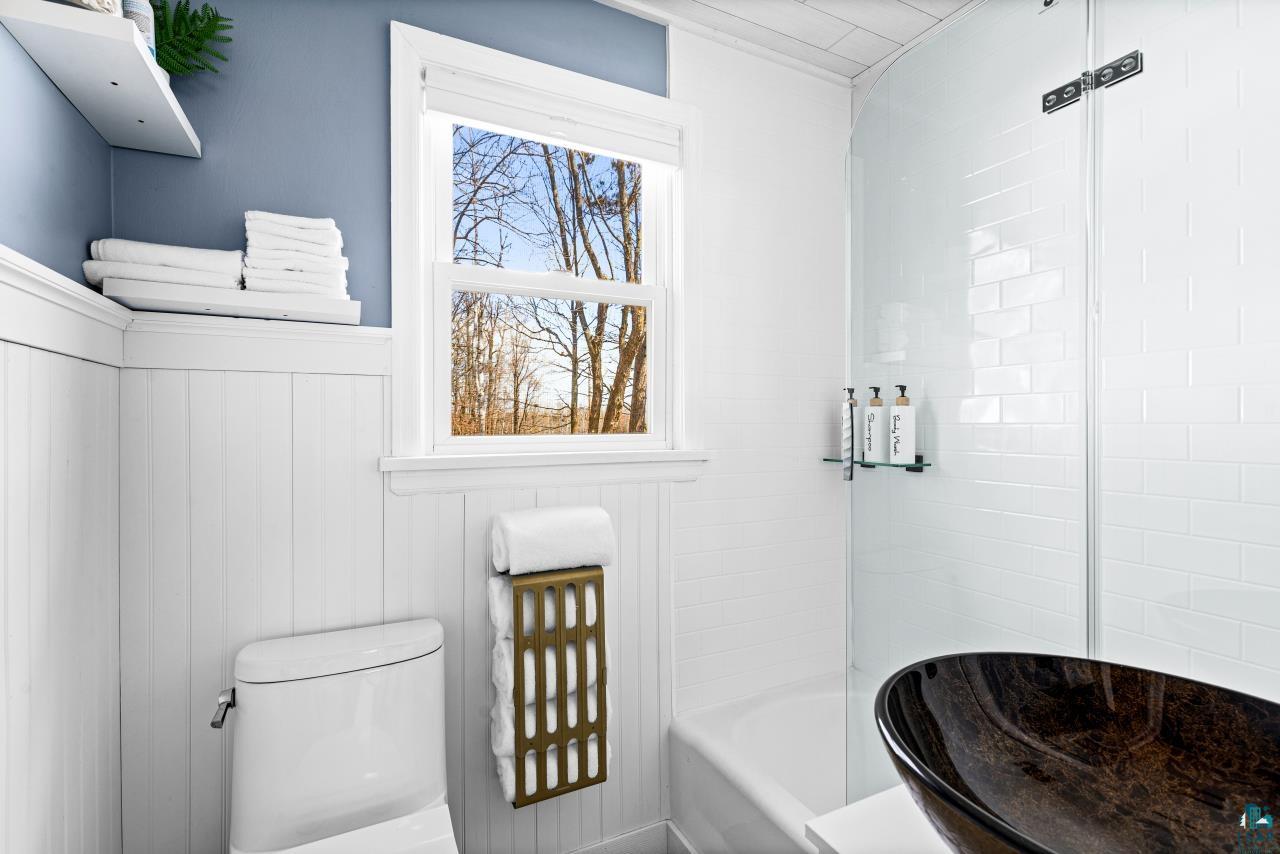 3645 Haines Road Hermantown, MN 55811 - Photo 33 of 35 Bathroom featuring wainscoting, a standard toilet, a window with outdoor scenery, a glass-enclosed shower with subway tile, and a vessel sink
