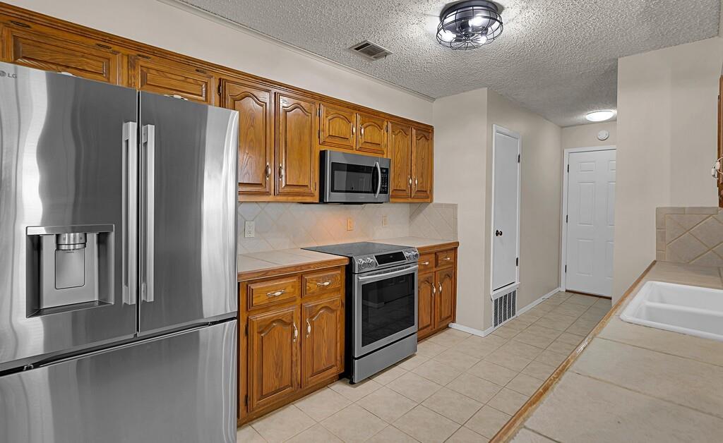2857 Country Spring Road Lorena, TX 76655 - Photo 8 of 40 Kitchen with stainless steel appliances, light tile patterned floors, decorative backsplash, wood finish cabinets, and a textured ceiling