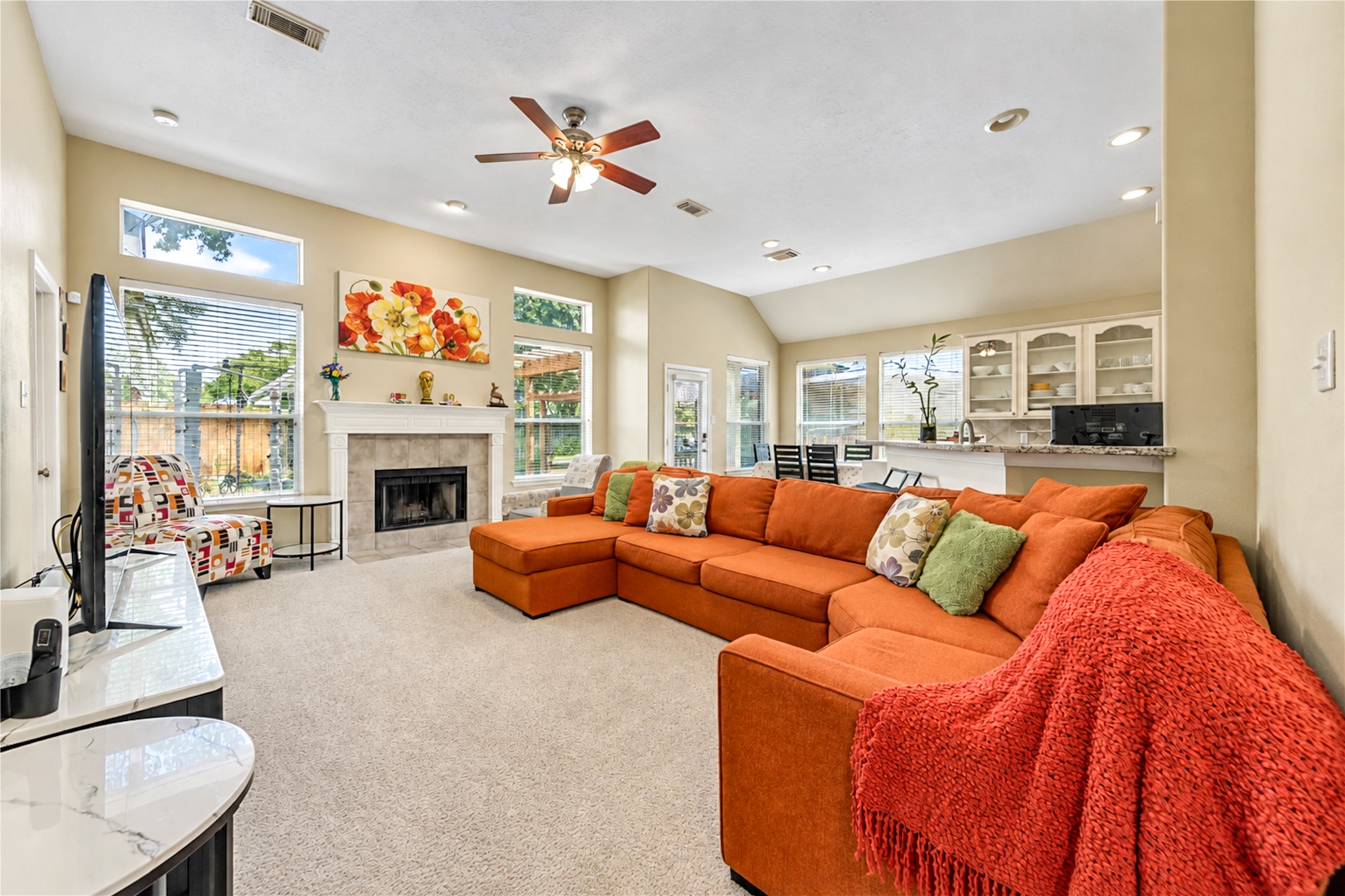 12806 Ridge Bank Lane Houston, TX 77041 - Photo 11 of 44 Spacious living room featuring a cozy fireplace, plush orange sectional, and abundant natural light, seamlessly flowing into a bright dining area and kitchen with built-in cabinetry.