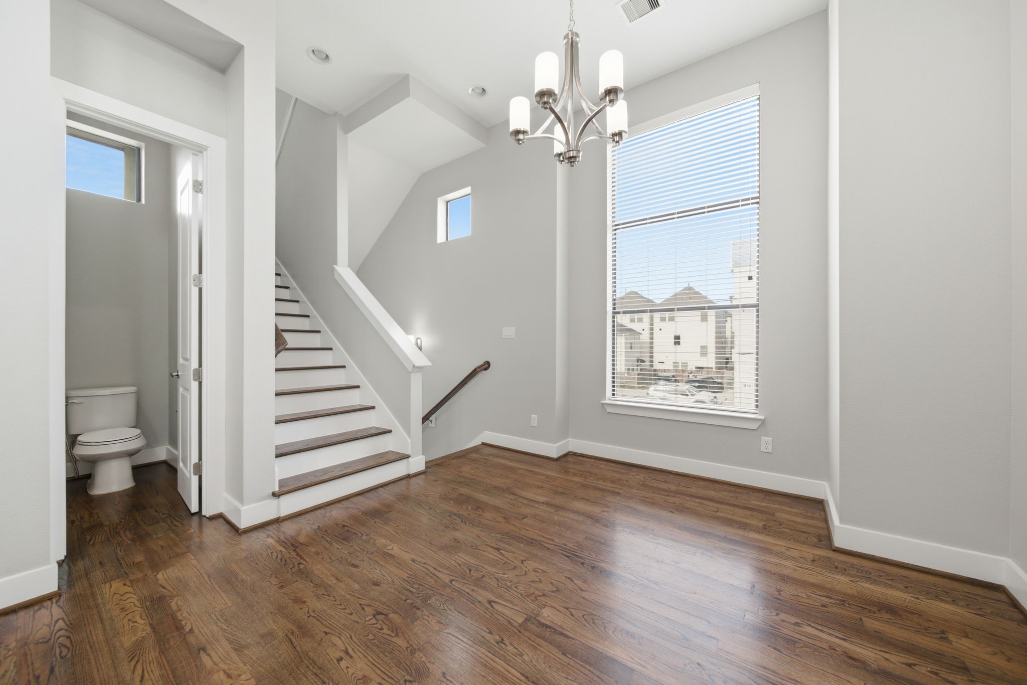 1814 Thompson Street Houston, TX 77007 - Photo 20 of 37 Bright, open-concept space featuring rich hardwood flooring, a modern chandelier, and expansive windows that fill the area with natural light. A stylish staircase and convenient powder bath complement the neutral color palette, creating a versatile and inviting space ideal for modern living and entertaining.