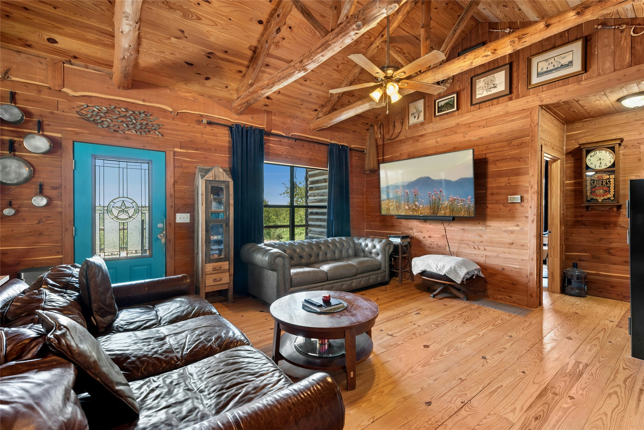 496 Middleton Road Spicewood, TX 78669 - Photo 9 of 40 Spacious interior featuring exposed wood-beam ceilings, wood-paneled walls, wood-finish flooring, a ceiling fan with integrated lighting, and a prominent turquoise entry door with a decorative glass insert