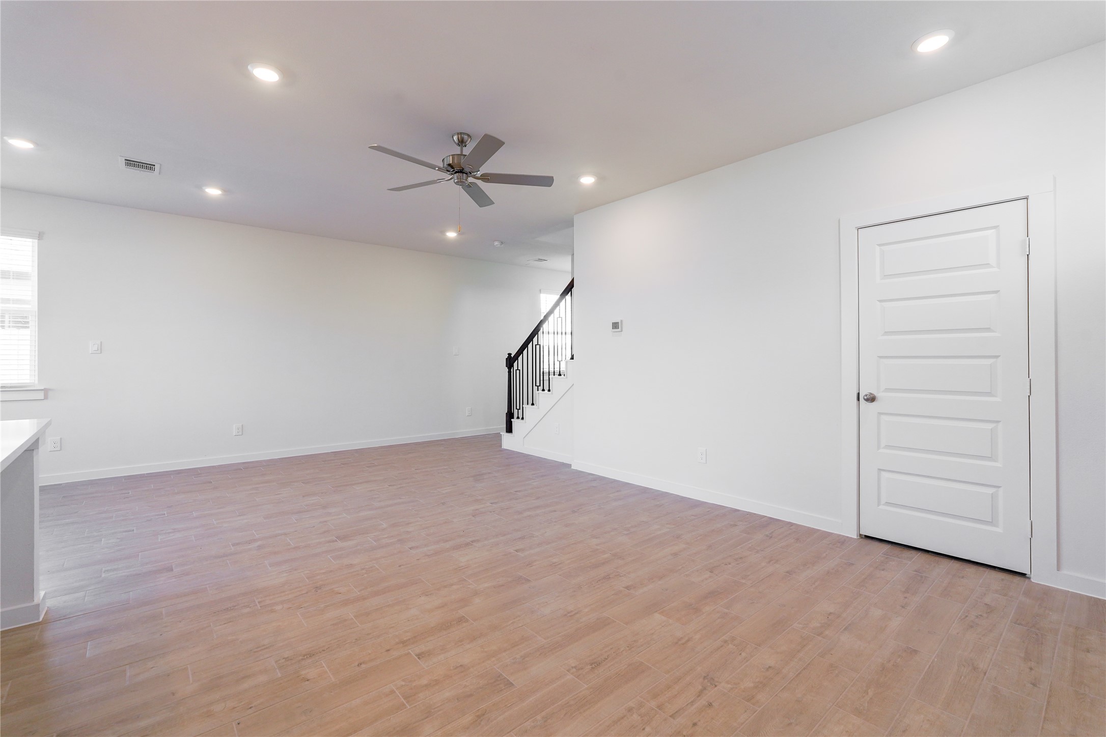 6618 Escondido Drive Rosharon, TX 77583 - Photo 11 of 31 This photo shows a spacious, open-concept room with light wood flooring and neutral walls. It features recessed lighting, a ceiling fan, and a staircase with dark railings. A white door is present, enhancing the modern, clean design.