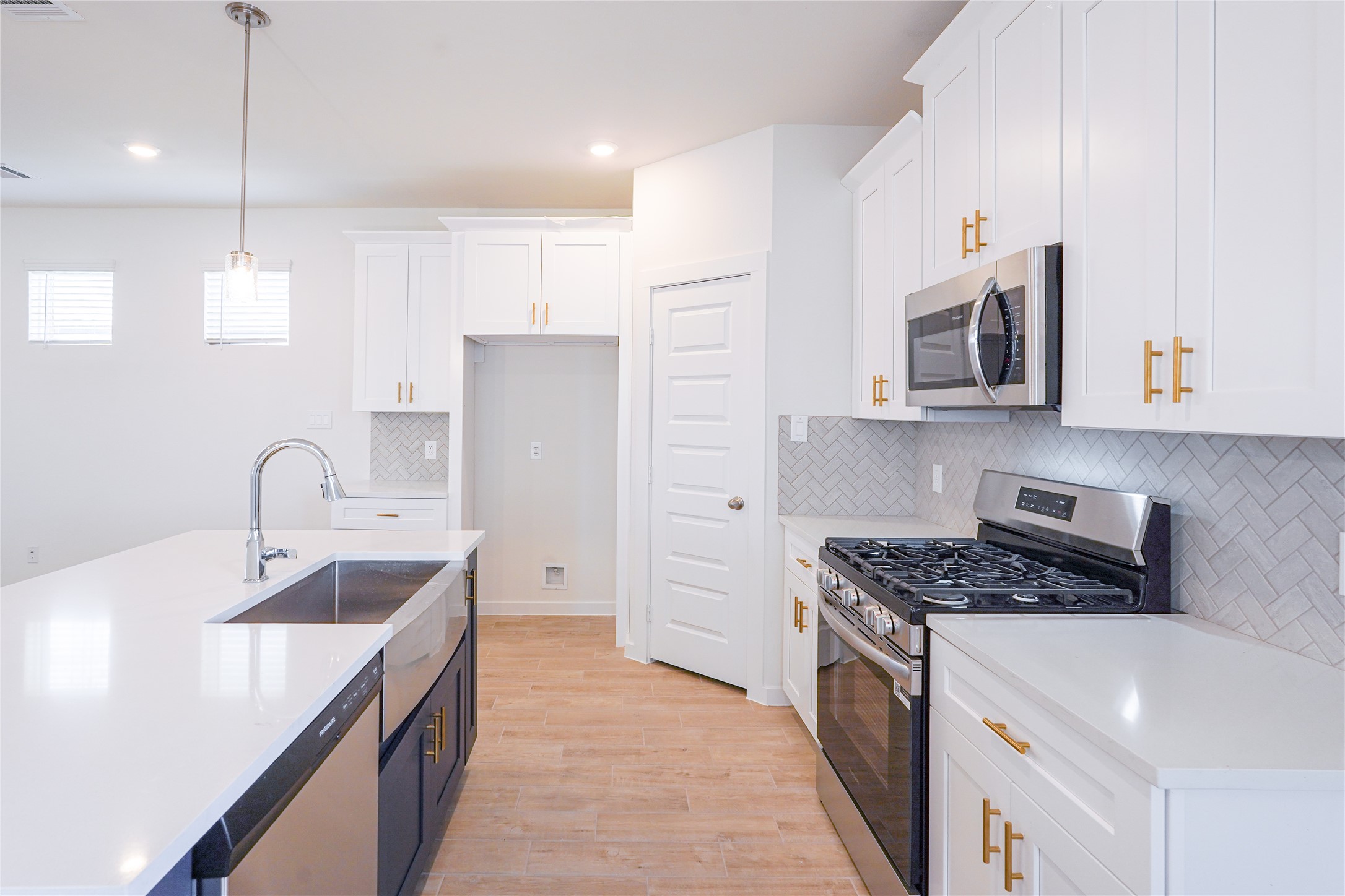 6618 Escondido Drive Rosharon, TX 77583 - Photo 14 of 31 Modern kitchen featuring sleek white cabinetry with gold hardware, stainless steel appliances, and a spacious island with a dual sink. The herringbone backsplash adds a stylish touch, complemented by light wood flooring and ample natural light.