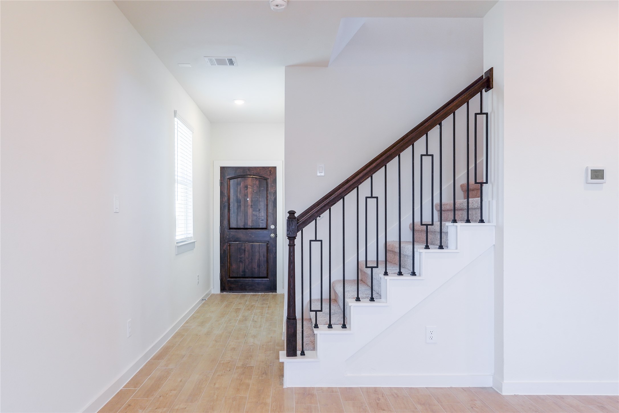 6618 Escondido Drive Rosharon, TX 77583 - Photo 5 of 31 This photo features a bright hallway with light wood flooring and a staircase with elegant dark wood handrails and black metal balusters. A window provides natural light, and a dark wooden door adds a touch of sophistication.