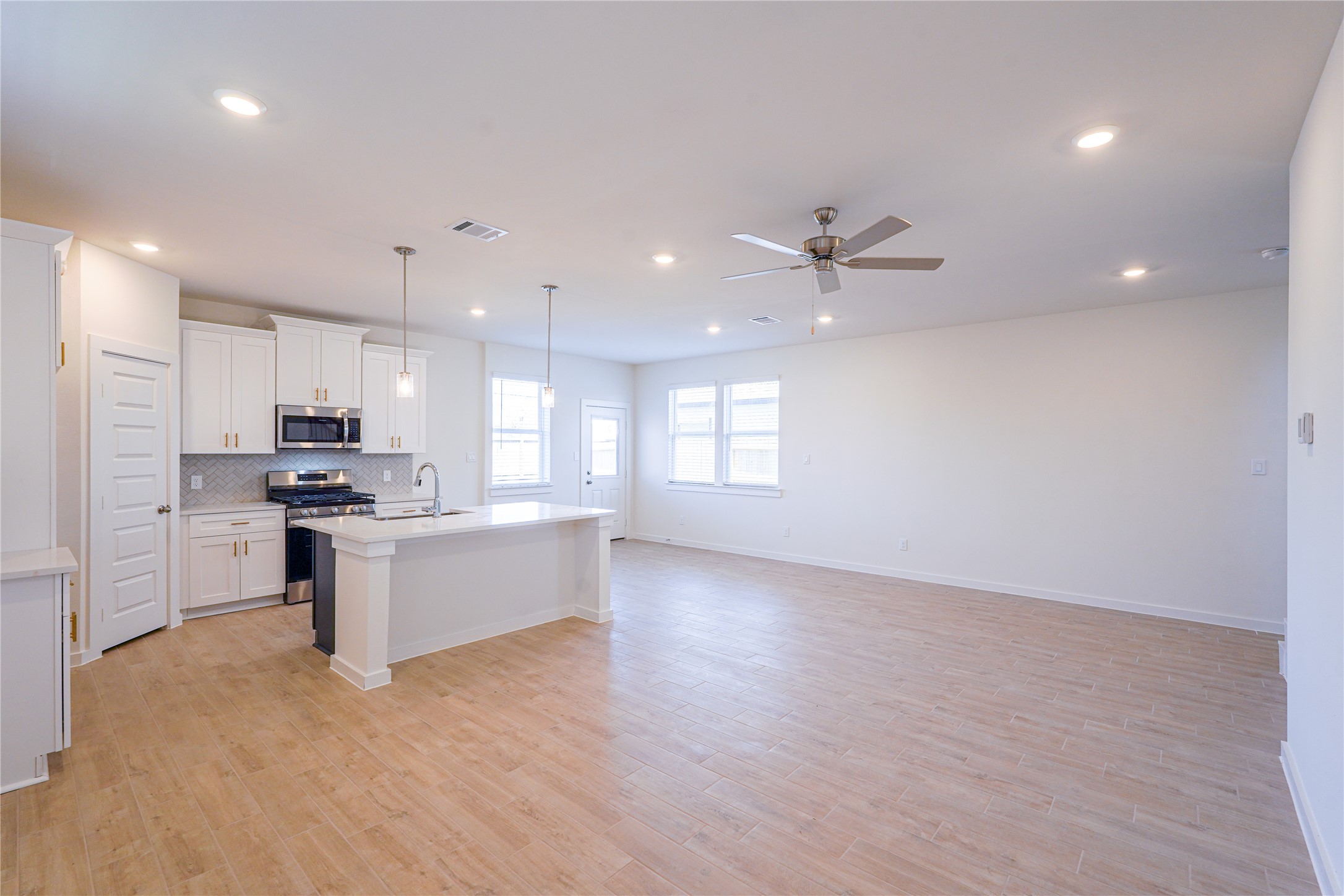 6618 Escondido Drive Rosharon, TX 77583 - Photo 8 of 31 This photo showcases a spacious open-concept living area with light wood flooring and neutral walls. The modern kitchen features white cabinetry, stainless steel appliances, and a central island with pendant lighting. The room is brightened by large windows and recessed lighting, and a ceiling fan adds comfort.