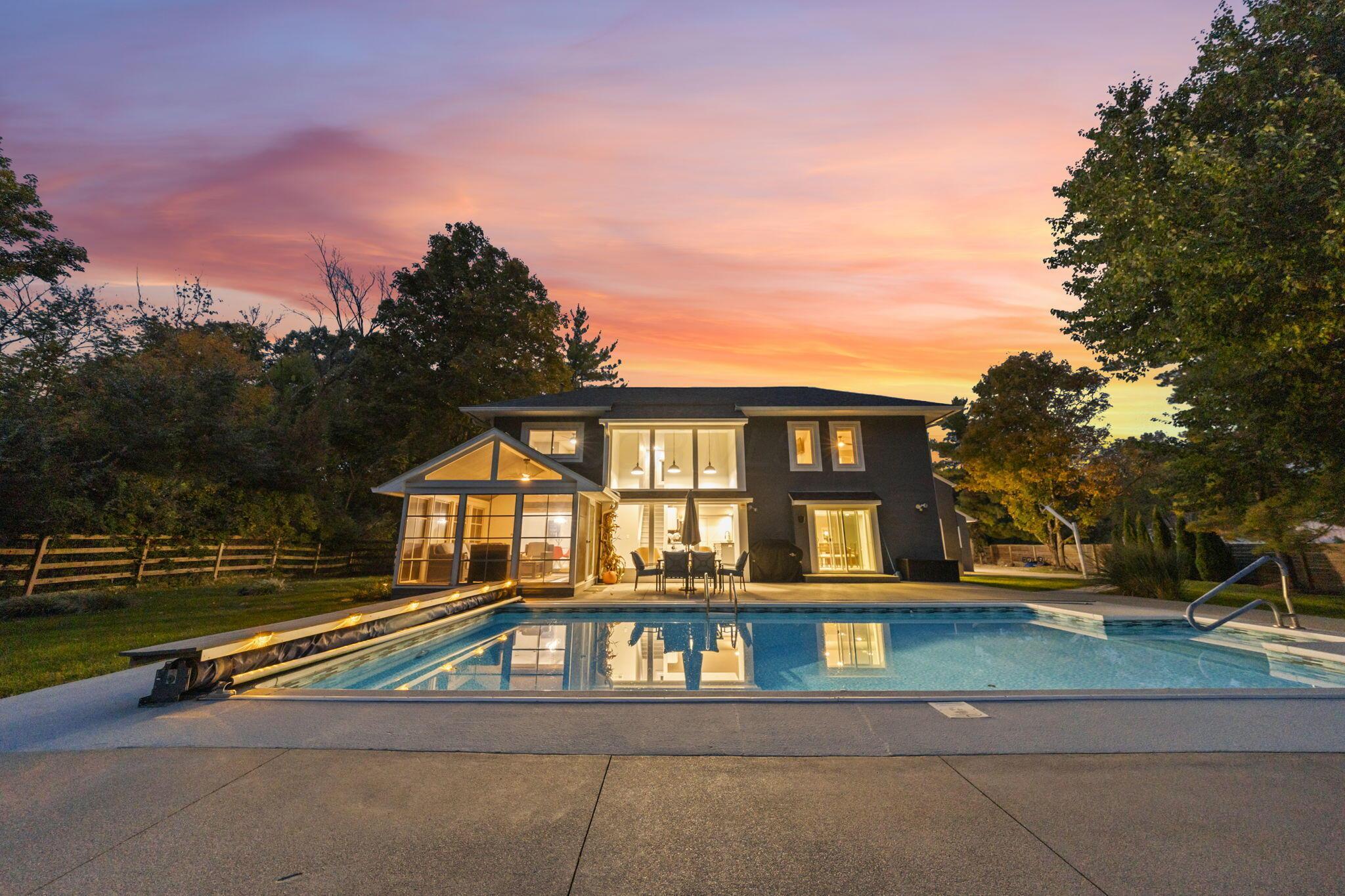 7140 Cook Road Powell, OH 43065 - Photo 8 of 87 Modern residence featuring a geometric facade with mixed material cladding, expansive glazing throughout the front elevation, a resort-style saltwater swimming pool with concrete decking, manicured lawn with mature landscaping, and ambient exterior lighting that illuminates the home at dusk