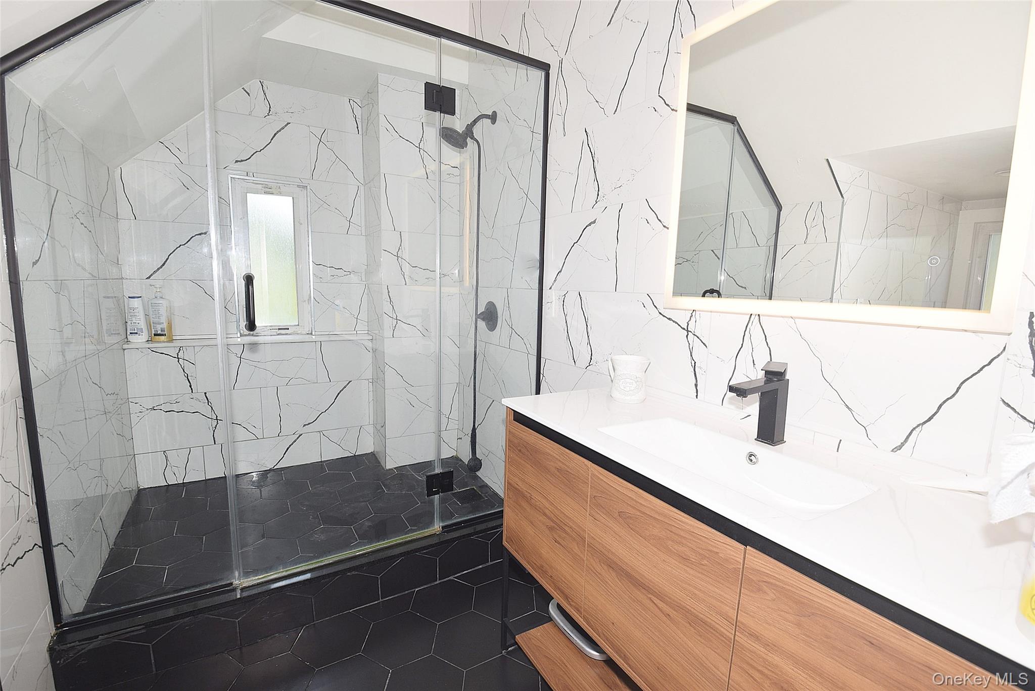 25 Old Mill Road Great Neck, NY 11023 - Photo 14 of 45 Frameless glass shower enclosure with black trim, a wood-finish vanity with an integrated sink, black hexagonal floor tiles, and a wall-mounted lighted mirror