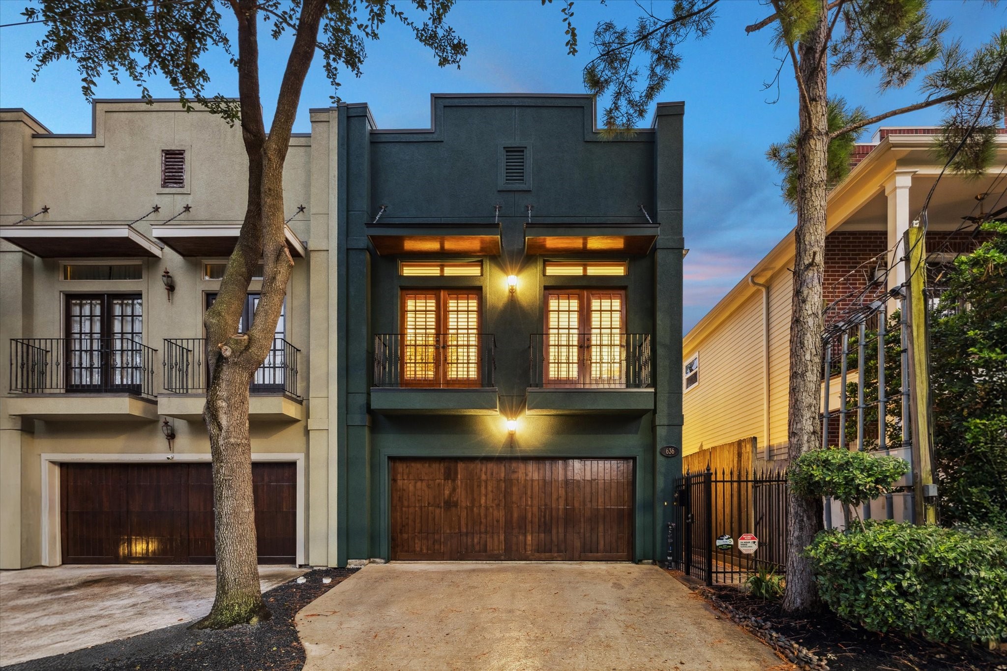 This modern townhouse features a striking dark green facade with warm wood accents. It includes a two-car garage, elegant balcony doors, and is framed by mature trees, offering a sophisticated urban living experience.