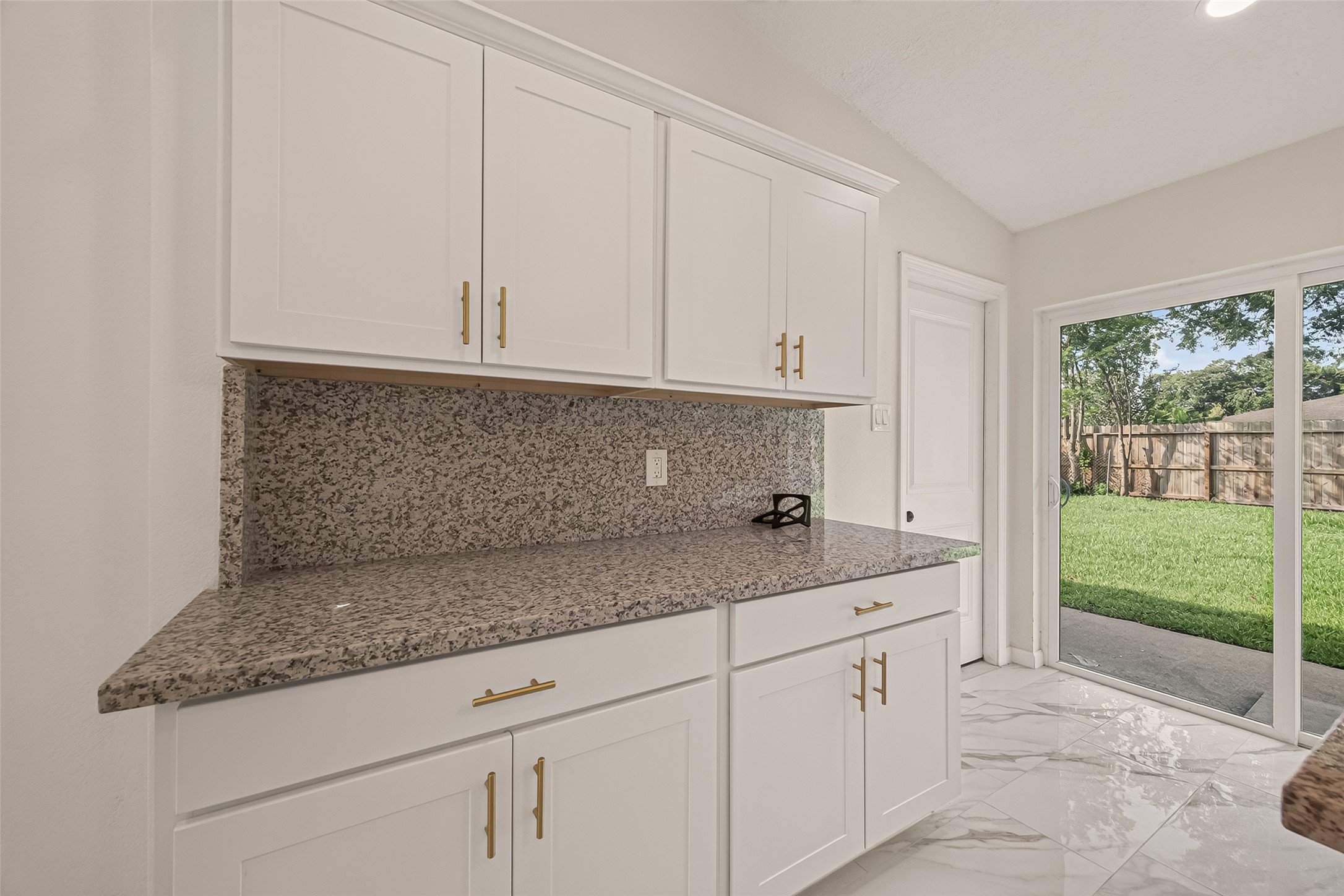 16619 Tibet Road Friendswood, TX 77546 - Photo 12 of 29 This photo showcases a bright kitchen space featuring elegant white cabinets with modern hardware, a speckled granite countertop, and a large sliding glass door opening to a lush, fenced backyard. The room is highlighted by glossy, tiled flooring.