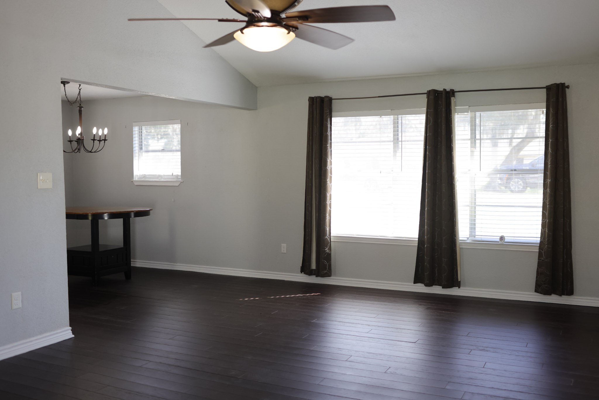 407 Tamarind Street Lake Jackson, TX 77566 - Photo 9 of 36 This photo shows a bright, open living space with dark wood flooring and large windows covered by curtains. There's a ceiling fan overhead, and a dining area with a chandelier visible. The room has a modern, neutral color scheme.