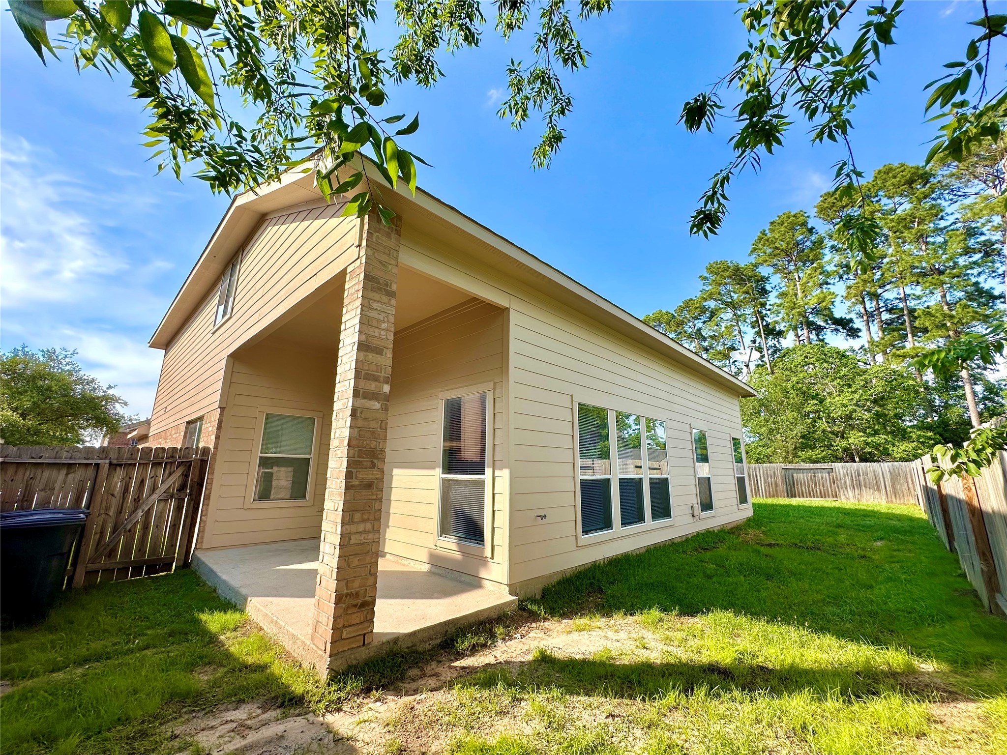 9611 Babbling Spring Court Tomball, TX 77375 - Photo 25 of 31 This photo shows a two-story house with beige siding and a brick pillar. It features a small covered patio area and large windows, surrounded by a fenced backyard with some trees, offering a pleasant outdoor space.