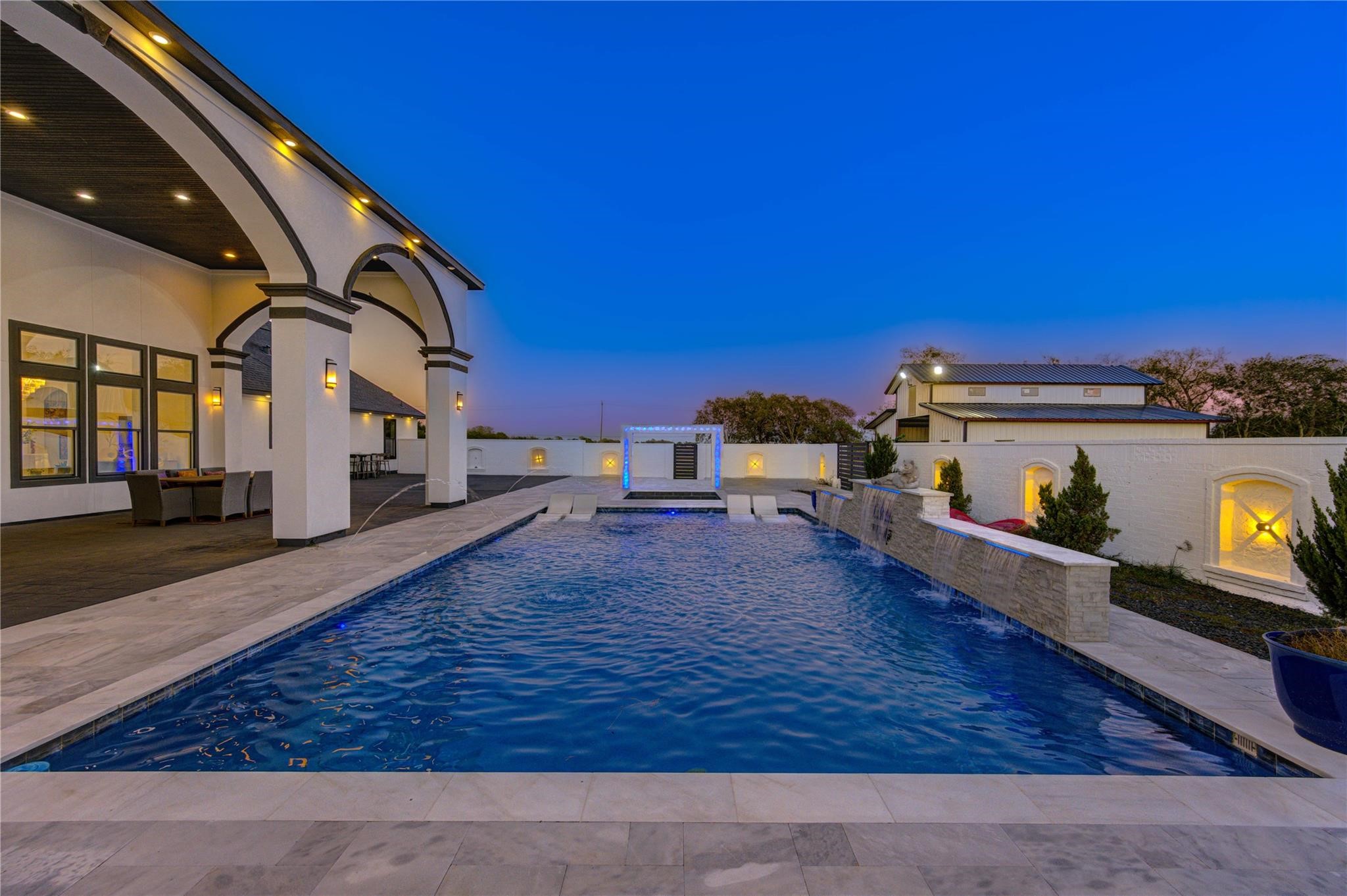 4821 Walters Road Richmond, TX 77469 - Photo 47 of 50 This stunning heated luxury pool creates a true resort-style experience with sleek modern design, expansive lounging areas, and breathtaking sunset views. Featuring elegant marble-style decking, in-water chaise loungers, ambient accent lighting, and a striking stone water feature, the space was thoughtfully designed for both relaxation and entertaining. A covered pergola lounge adds the perfect setting for evening gatherings, while the oversized pool offers a seamless blend of comfort & privacy.