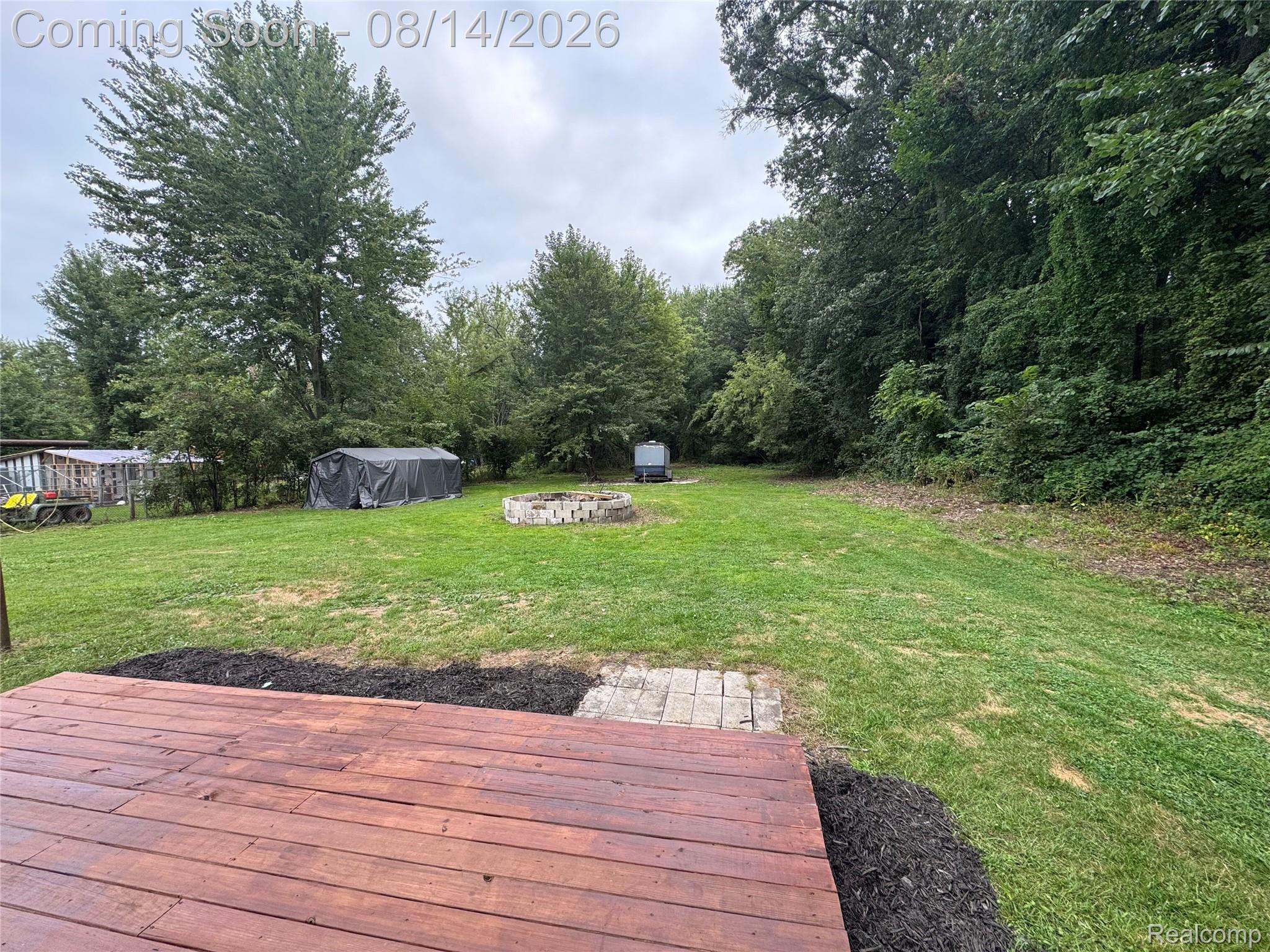 Wood-stained deck with dark mulch landscaping, expansive green lawn, stone fire pit, and mature trees providing natural privacy