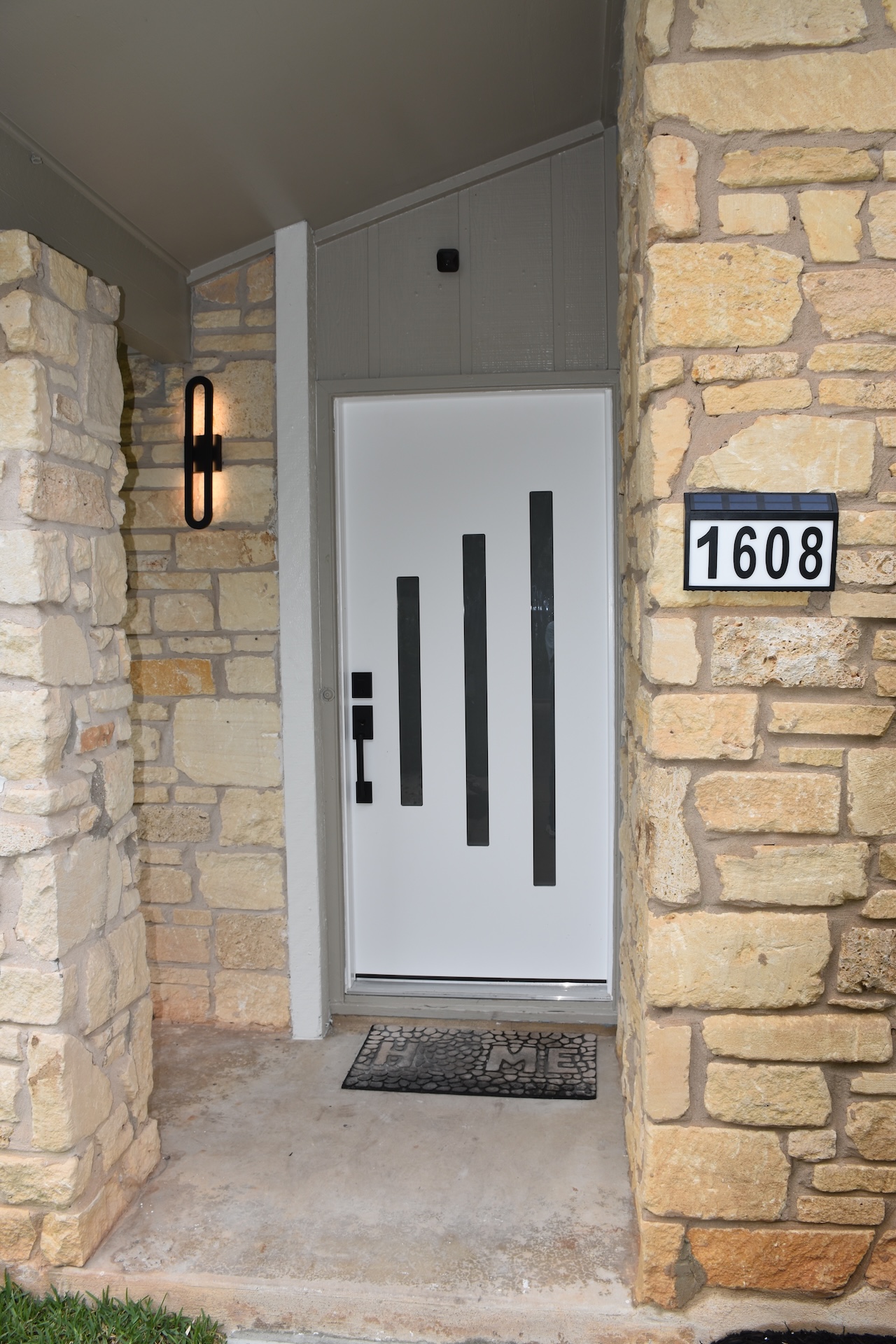 1608 Springer Lane Austin, TX 78758 - Photo 7 of 35 Modern entry featuring a white door with black vertical accents, complemented by stone facade pillars and a contemporary wall sconce