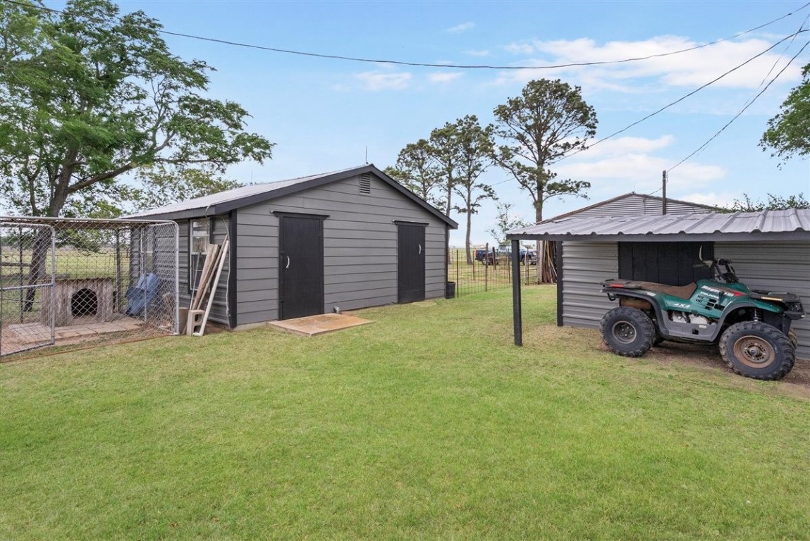 13339 Bj Dusek Road Wallis, TX 77485 - Photo 23 of 39 This photo shows a backyard with a well-maintained lawn, featuring two grey storage sheds. One shed has an attached fenced area, possibly for pets. There's also a covered area with an ATV parked underneath. The setting is surrounded by trees, providing a rural and spacious feel.