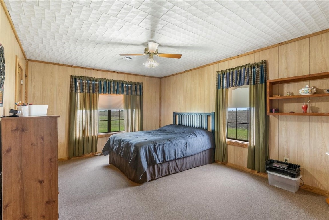 13339 Bj Dusek Road Wallis, TX 77485 - Photo 6 of 39 Cozy bedroom with wood-paneled walls and carpeted floor, Large windows with green curtains provide natural light and countryside views. Includes a ceiling fan, and built-in shelving.