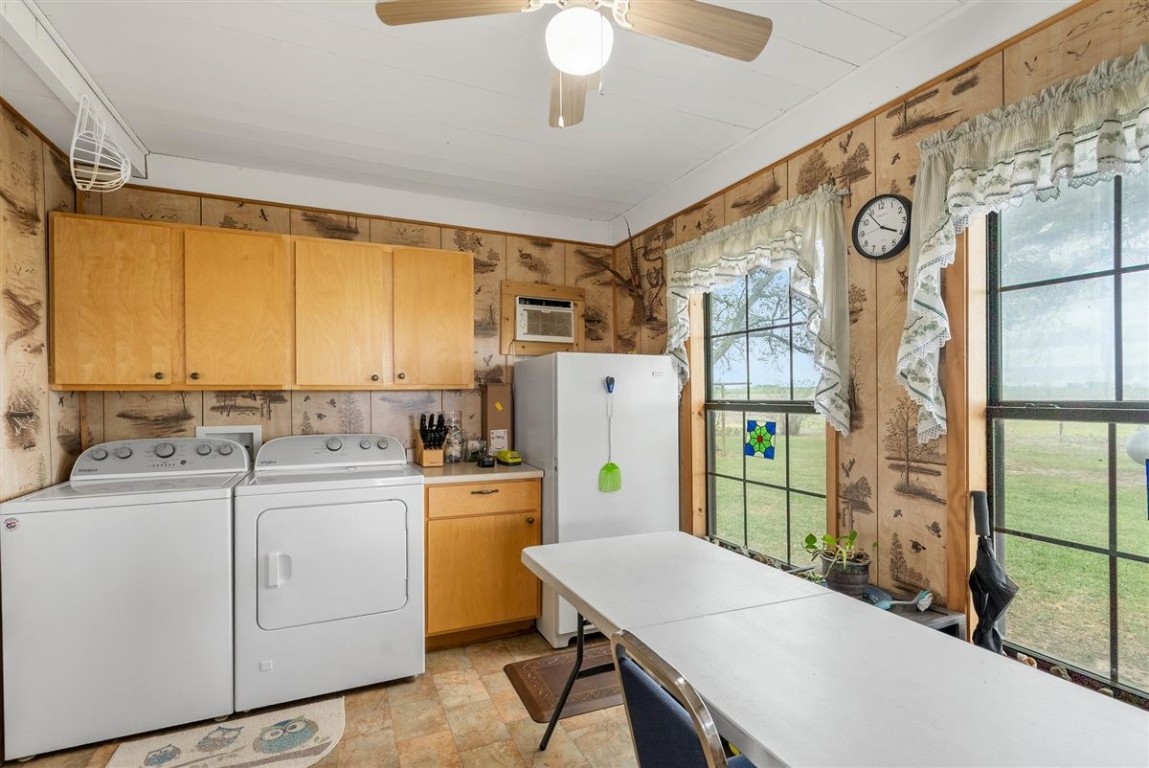 13339 Bj Dusek Road Wallis, TX 77485 - Photo 7 of 39 This photo showcases a cozy laundry room with a washer, dryer, and fridge. It features wooden cabinets, a ceiling fan, and large windows with countryside views, offering ample natural light.