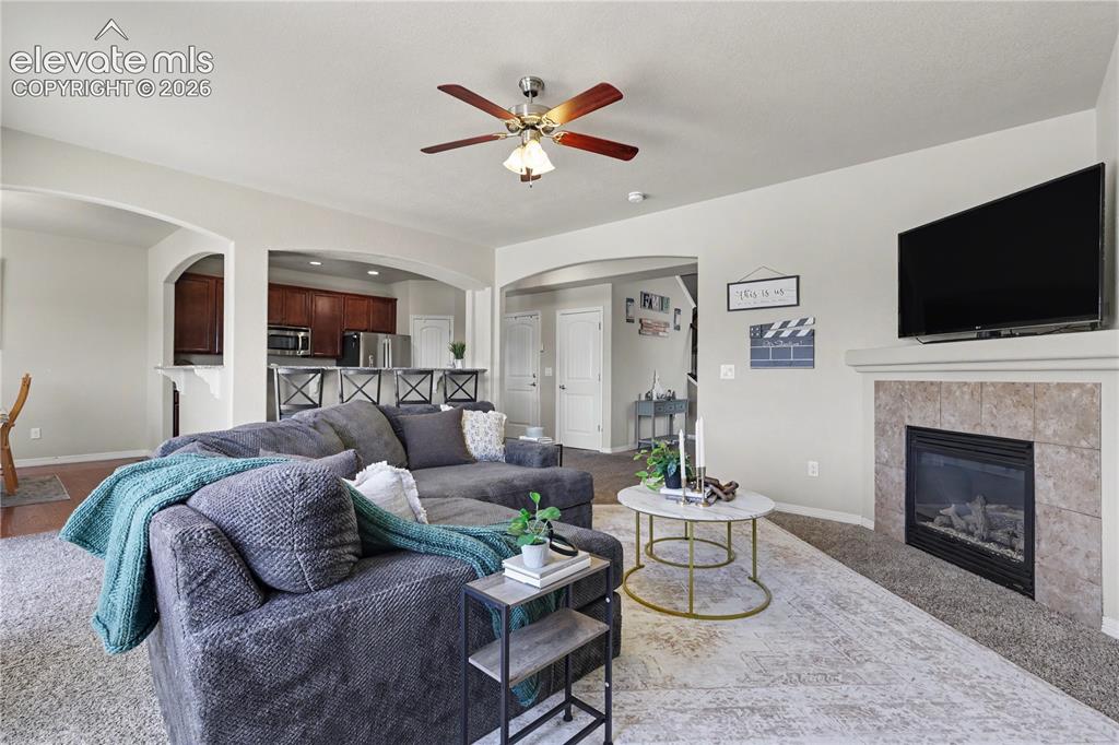6598 Alliance Loop Colorado Springs, CO 80925 - Photo 7 of 39 Living area featuring a gas fireplace with a tiled surround and mantel, overhead ceiling fan, and carpet flooring