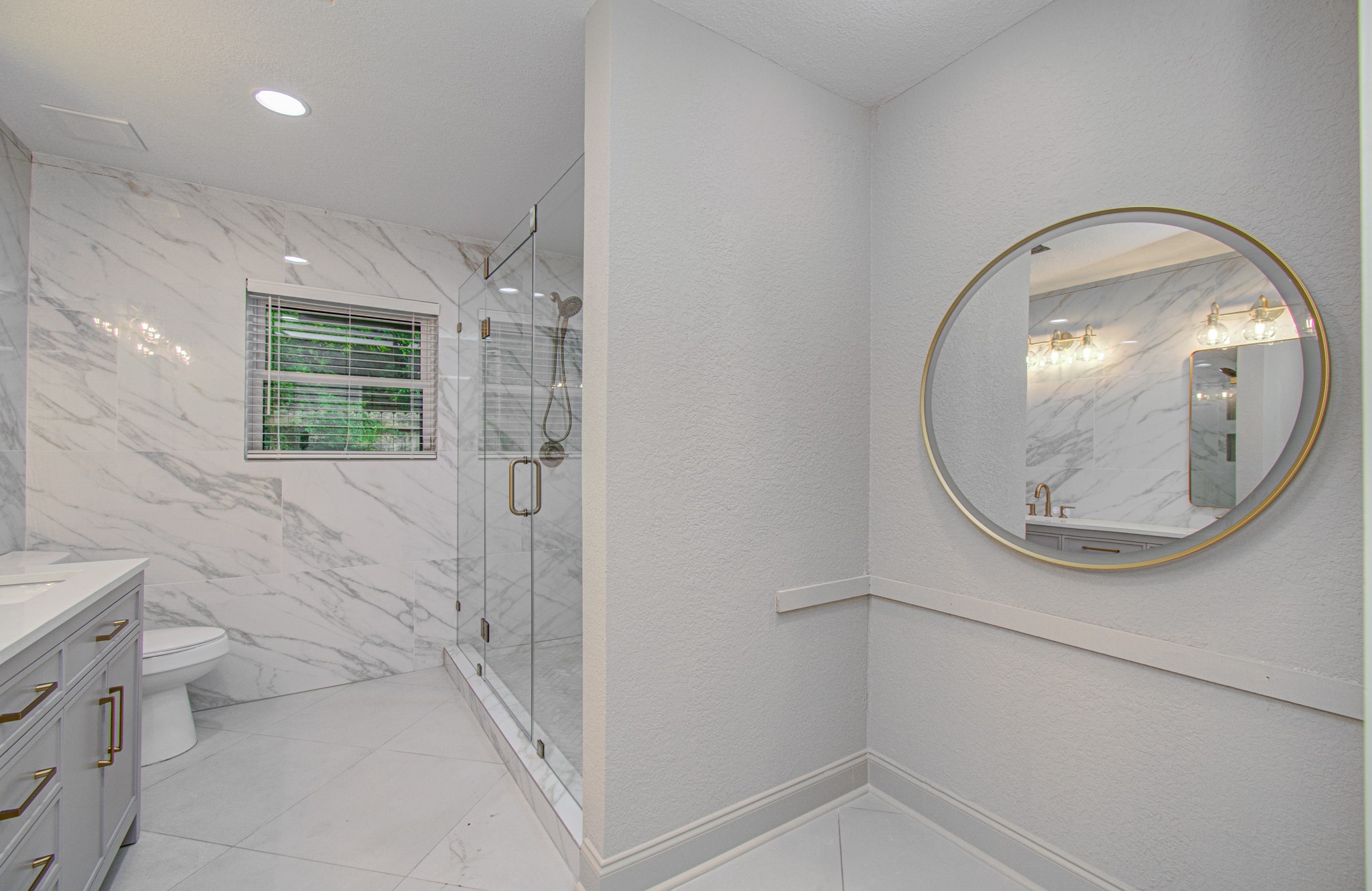1801 Benbow Way Houston, TX 77080 - Photo 21 of 32 This modern bathroom features a spacious glass-enclosed shower with marble-style tiles, a sleek vanity with ample storage, and a large round mirror. Natural light enters through a window with blinds, creating a bright and elegant space.