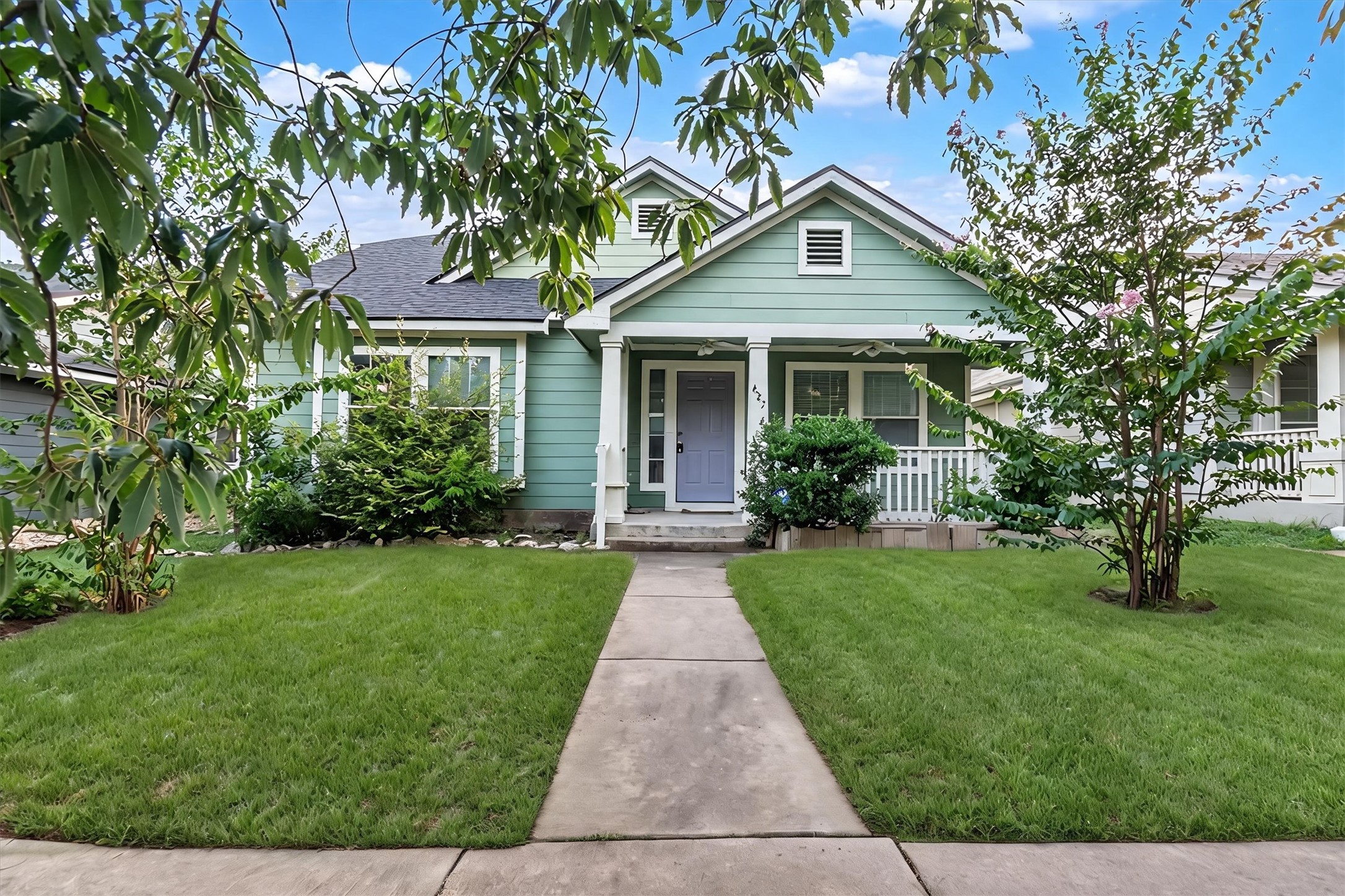 A charming sage green home with a welcoming front porch, flanked by mature landscaping and a well-kept lawn.