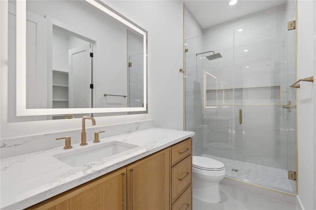 6038 Prospect Avenue Dallas, TX 75206 - Photo 37 of 40 Bathroom featuring a large vanity with an integrated sink and light wood-finish cabinetry, a lighted mirror, a glass-enclosed shower with a rain showerhead, and gold-tone fixtures
