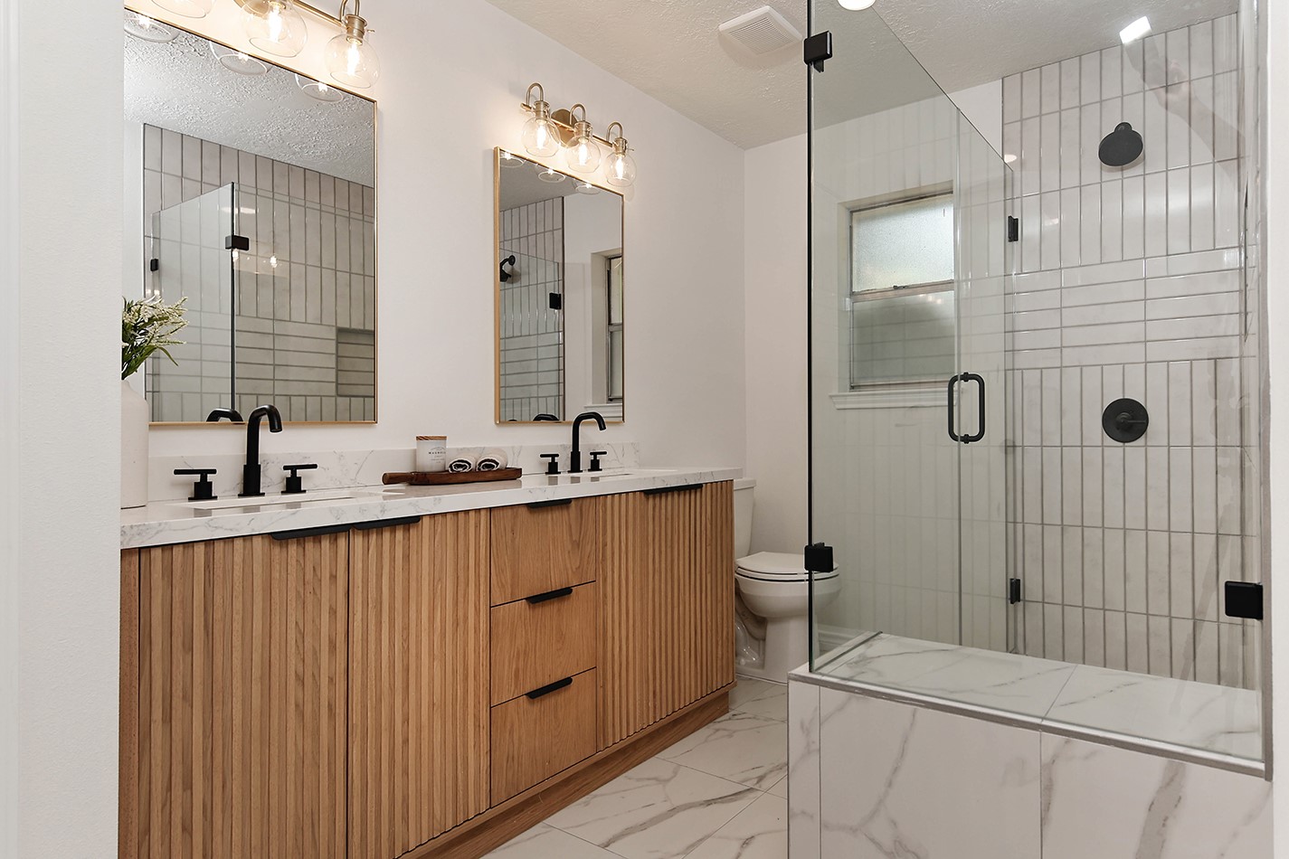 19410 Craigchester Lane Spring, TX 77388 - Photo 28 of 32 Stunning primary bath has custom wood vanity with reed design, accented with black pulls and convenient dual sinks in quartz and modern black plumbing fixtures. Gold light fixtures with gold mirrors add same warmth as the cabinet's wood color. Linen storage and walk-in closet are accessed on the opposite side of bathroom.