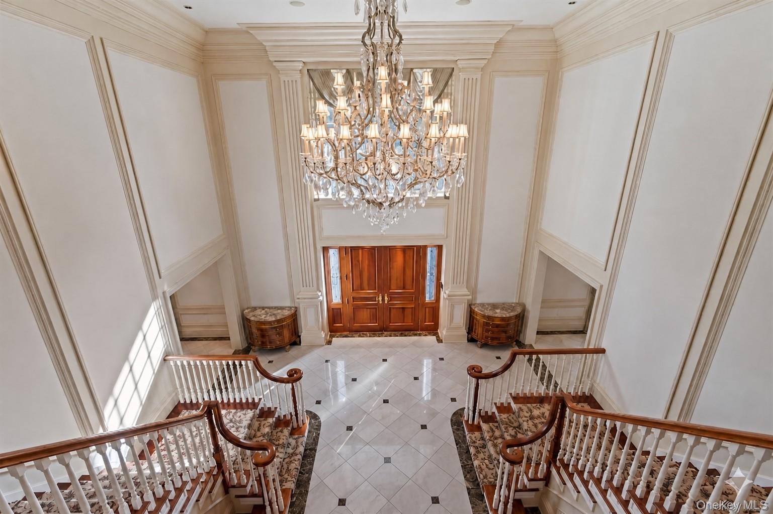 660 Chicken Valley Road Locust Valley, NY 11560 - Photo 17 of 48 Grand two-story foyer featuring a large crystal chandelier, a double wooden entry door, a marble tile floor with dark accents, and twin curved staircases with patterned runners