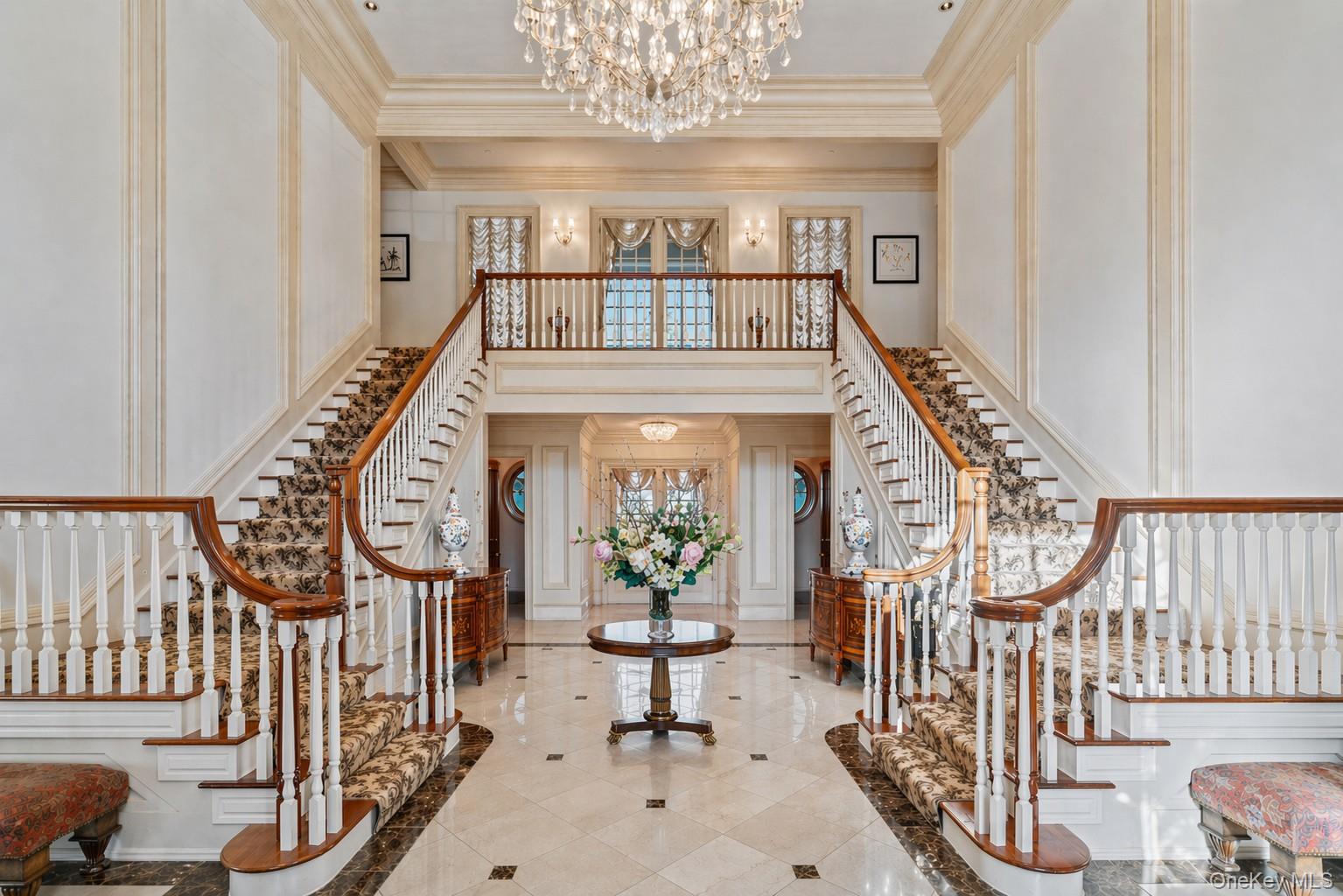 660 Chicken Valley Road Locust Valley, NY 11560 - Photo 3 of 48 Grand foyer featuring a dual staircase with patterned runners, a sparkling crystal chandelier, wall paneling, and a polished tile floor with dark accents
